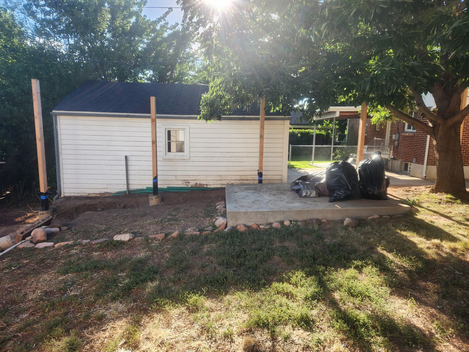 Small white shed in a sunny backyard with trees, a porch, and trash bags beside it.