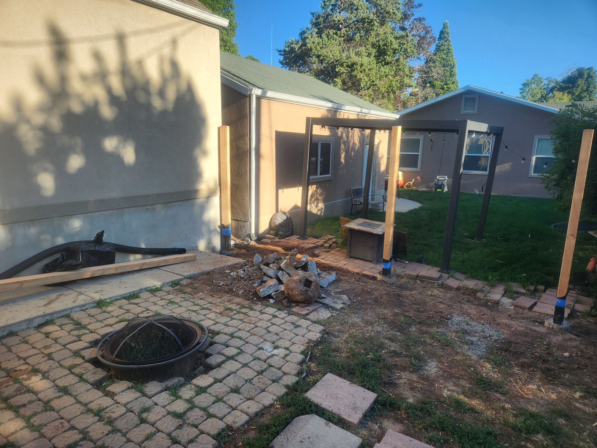 Backyard patio with broken pavers, scattered debris, and a fire pit near a house and fence