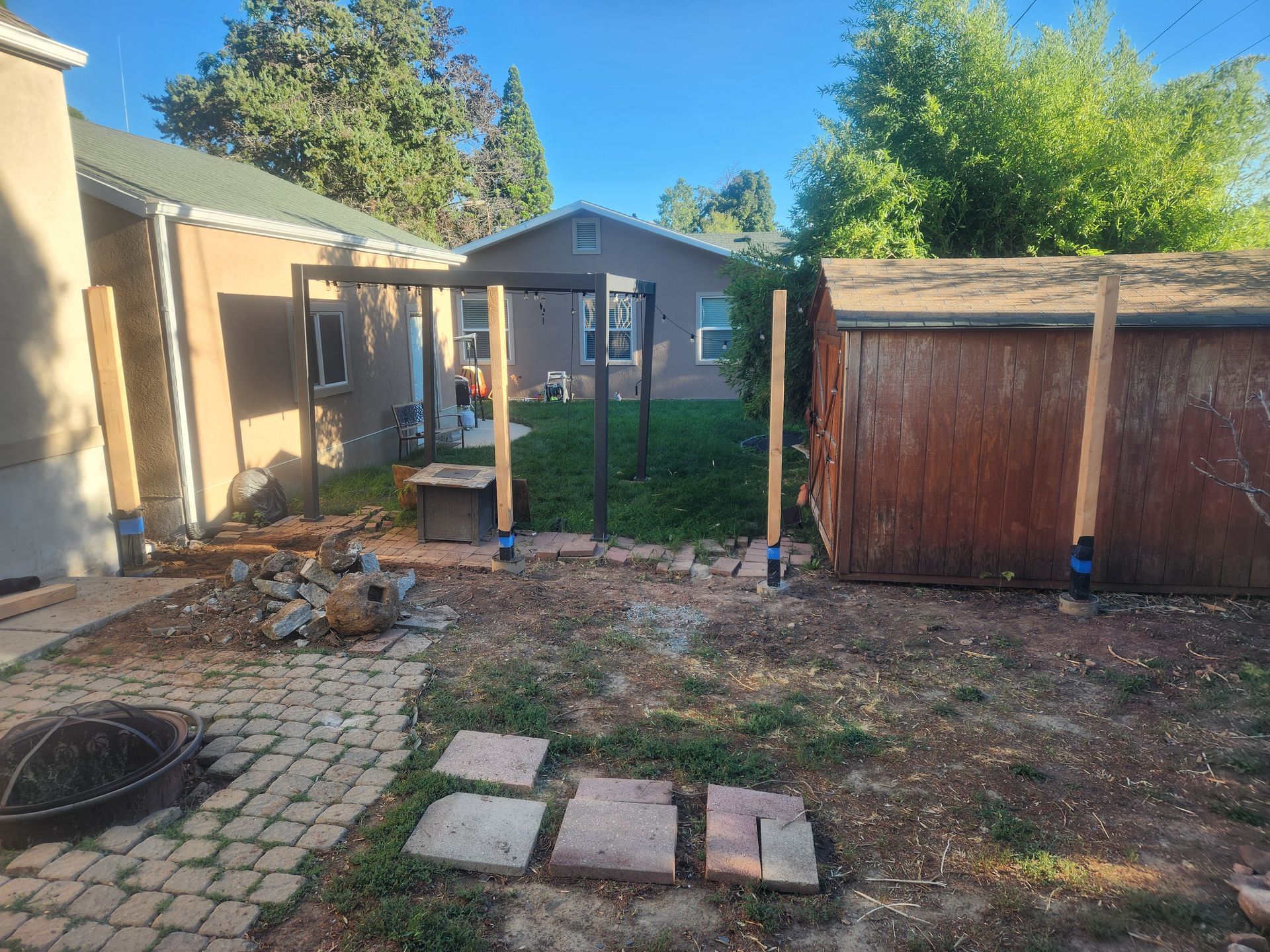Backyard with patio stones, dirt yard, wooden shed, and a small gray house under trees.