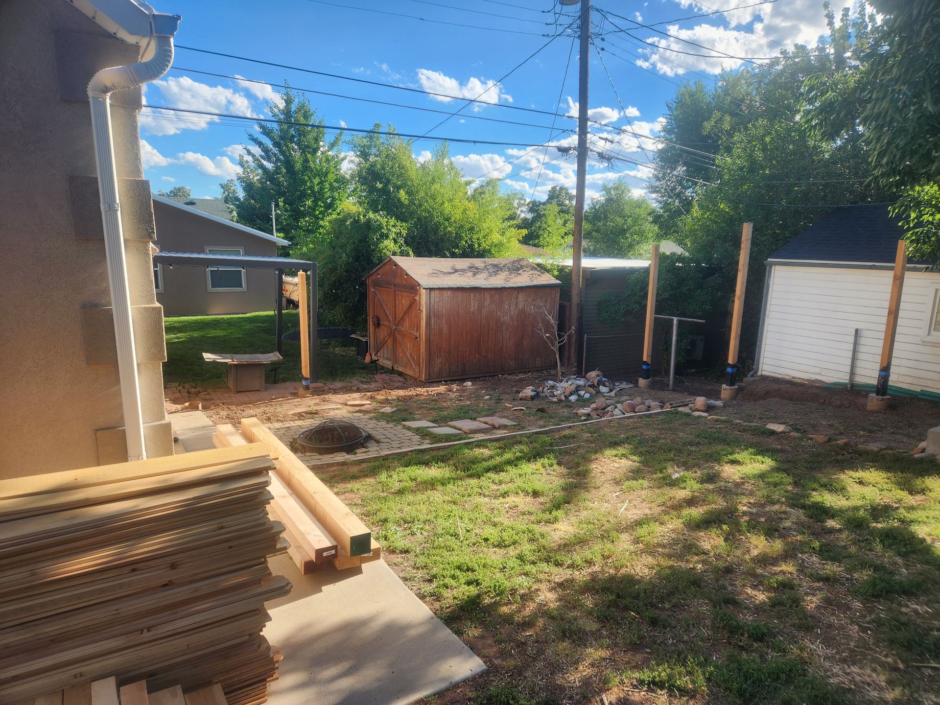 Backyard with stacked lumber, a small shed, and a white garage under a bright blue sky