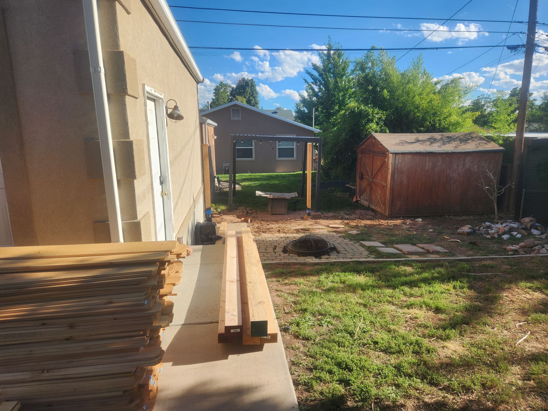 Backyard with stacked lumber on a concrete slab, dirt patches, a small shed, and a house under a blue sky.
