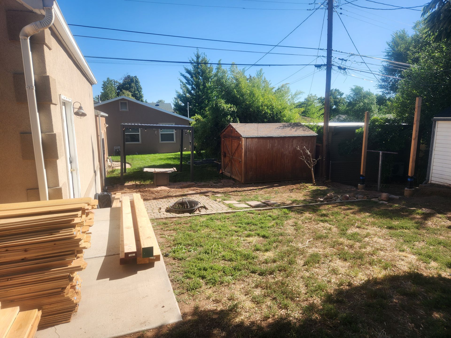 Backyard with patchy grass, stacked lumber, a small shed, and neighboring houses under a blue sky