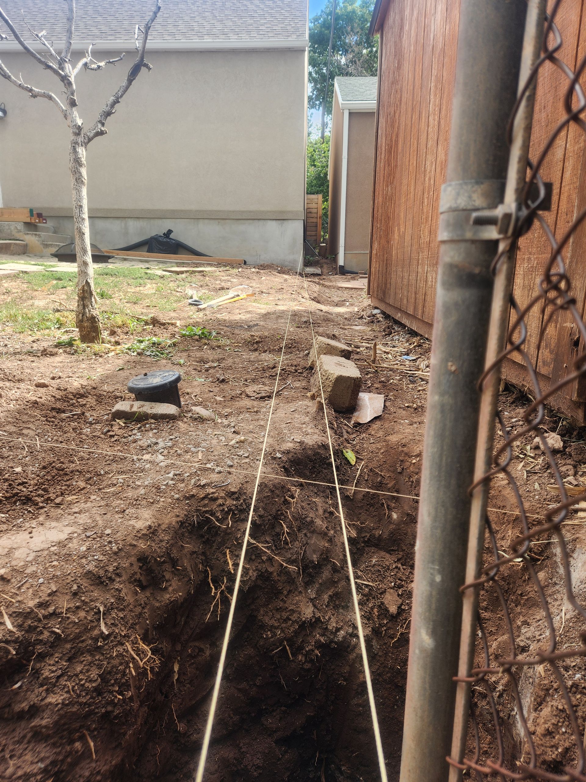 Backyard with a large trench beside a fence and house, dirt piled around it.
