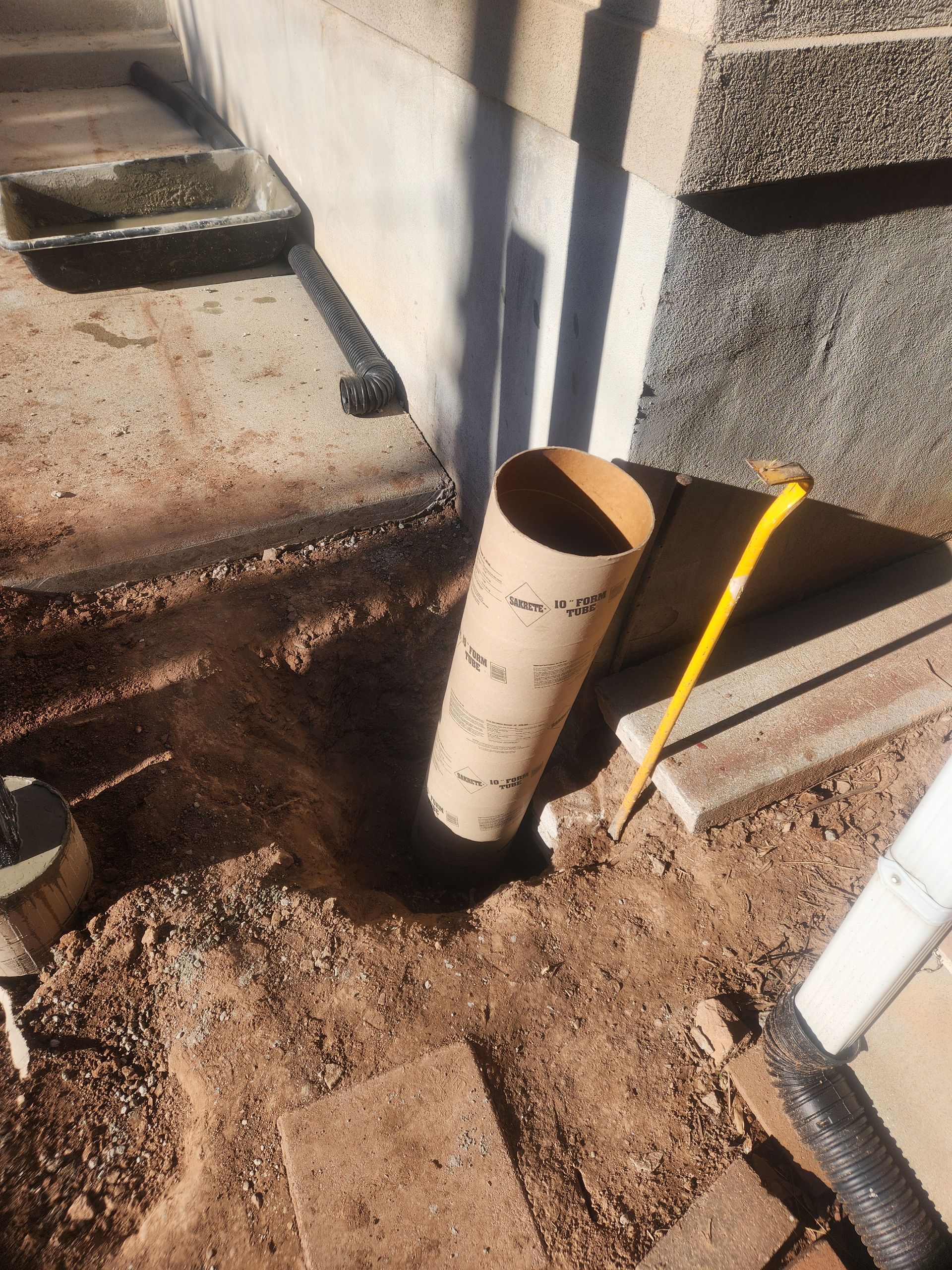 Open PVC pipe beside a concrete wall, with dirt and a yellow-handled tool nearby.