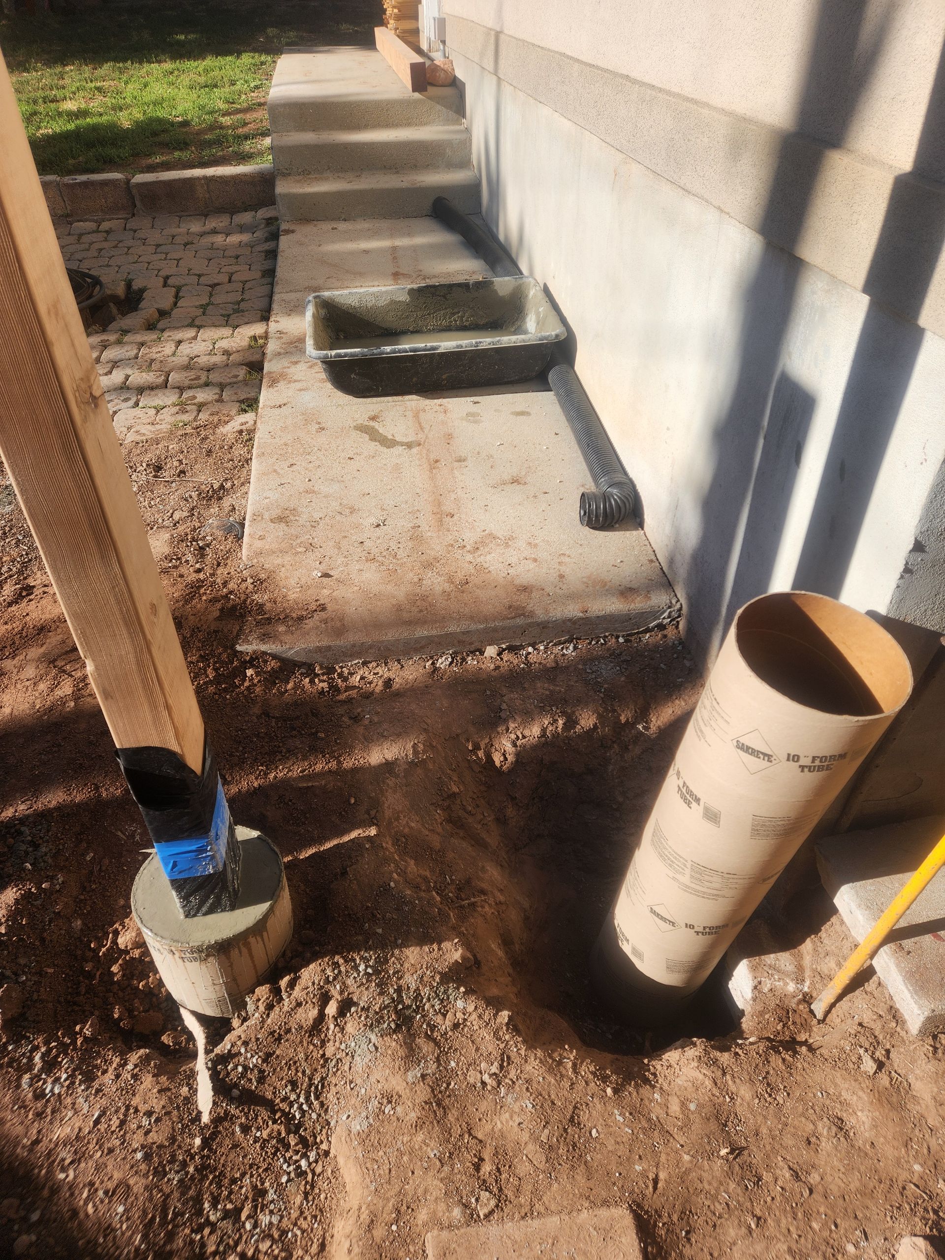 Concrete walkway under construction beside a house, with dirt, a utility pipe, and a small bucket.
