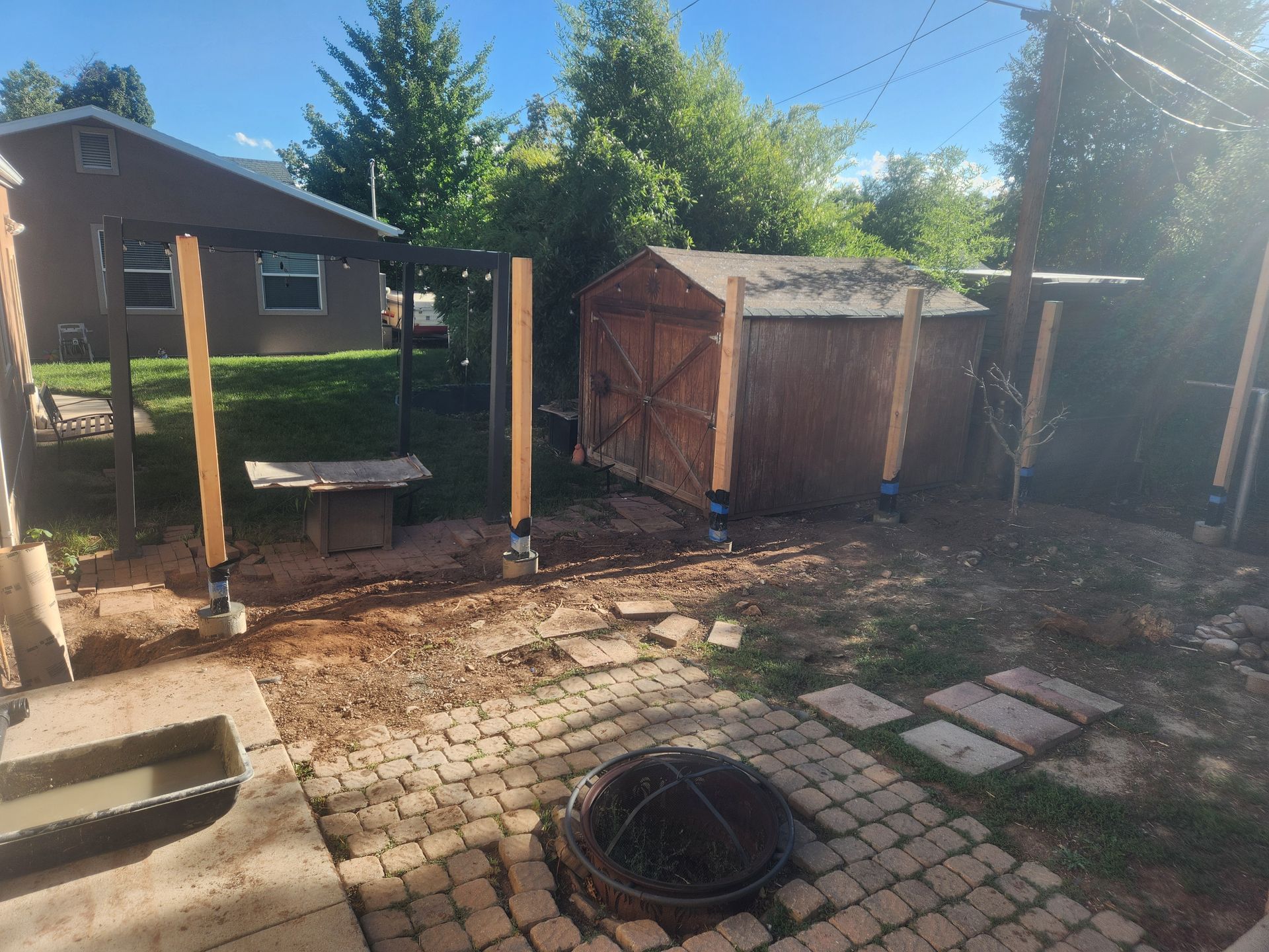 Backyard patio with brick pavers, wooden posts, a shed, and a fire pit under sunny sky
