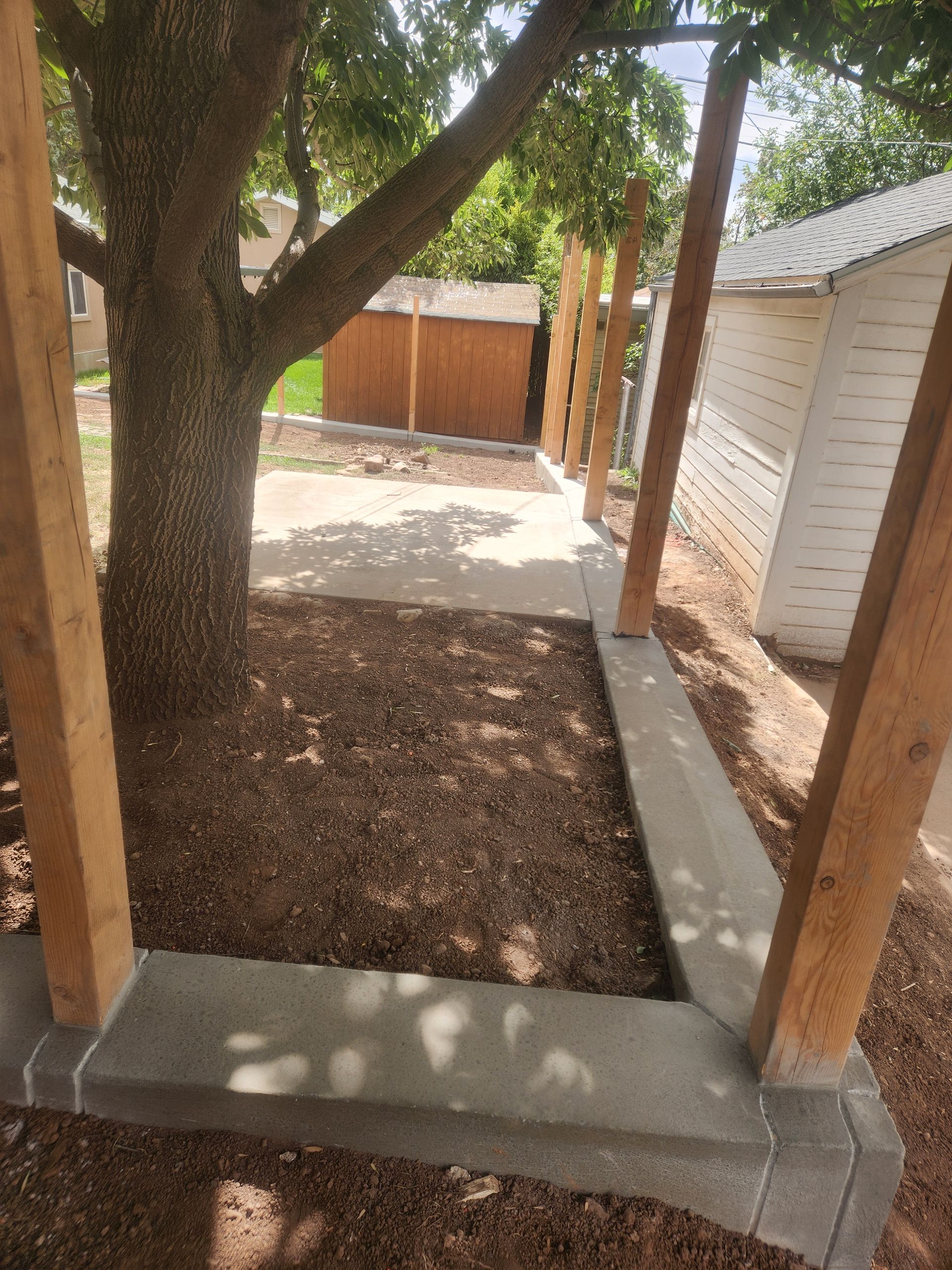 Narrow backyard walkway with trees, wooden posts, gravel, and a concrete border beside a shed
