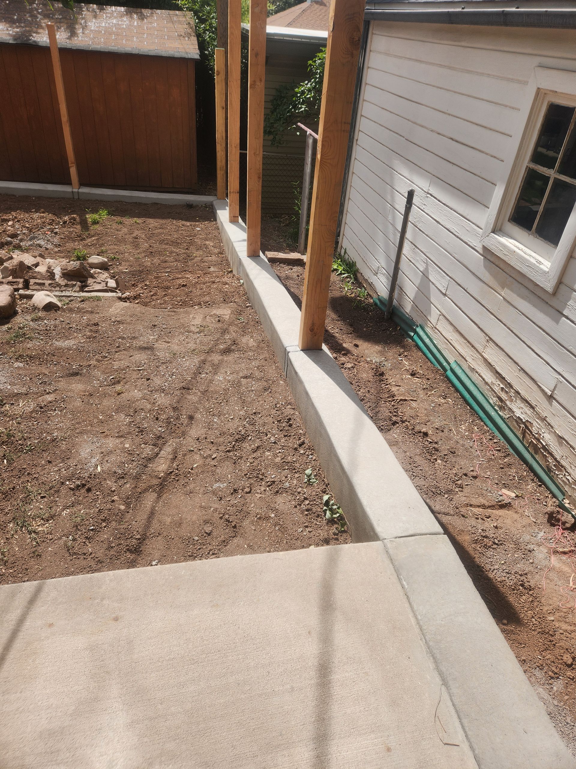 Narrow concrete walkway beside a house, leading past a gravel yard and wooden posts to a fenced area.