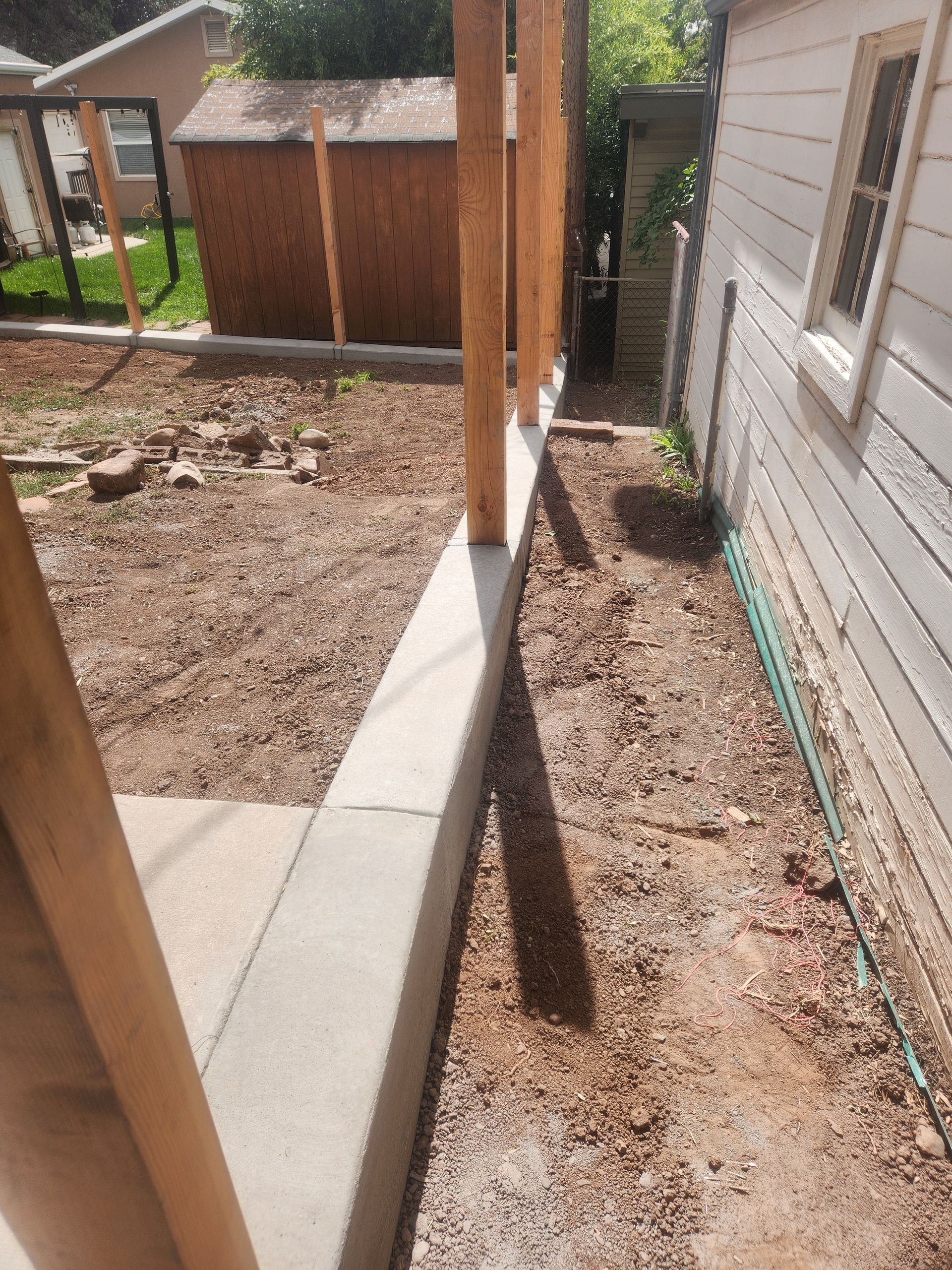 Narrow outdoor side yard with gravel, a concrete path, wooden posts, and a white house wall in sunlight