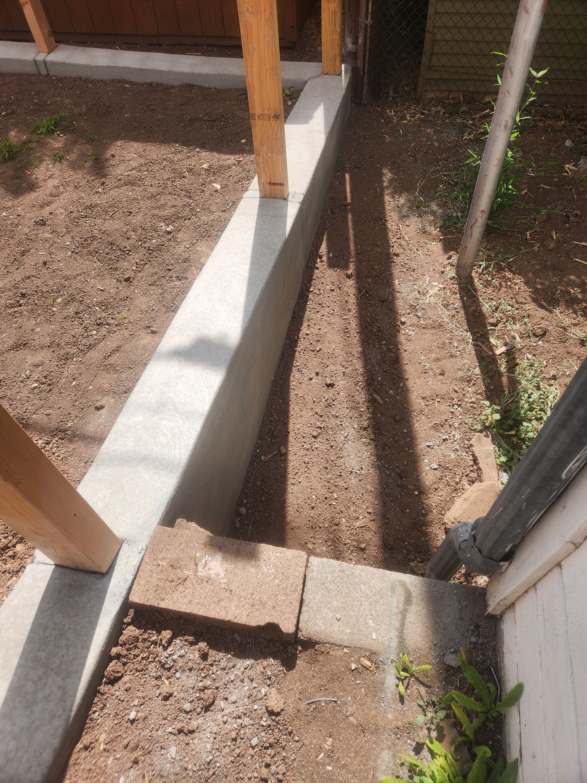 Narrow gravel trench beside a house foundation with wooden posts and concrete footing under construction