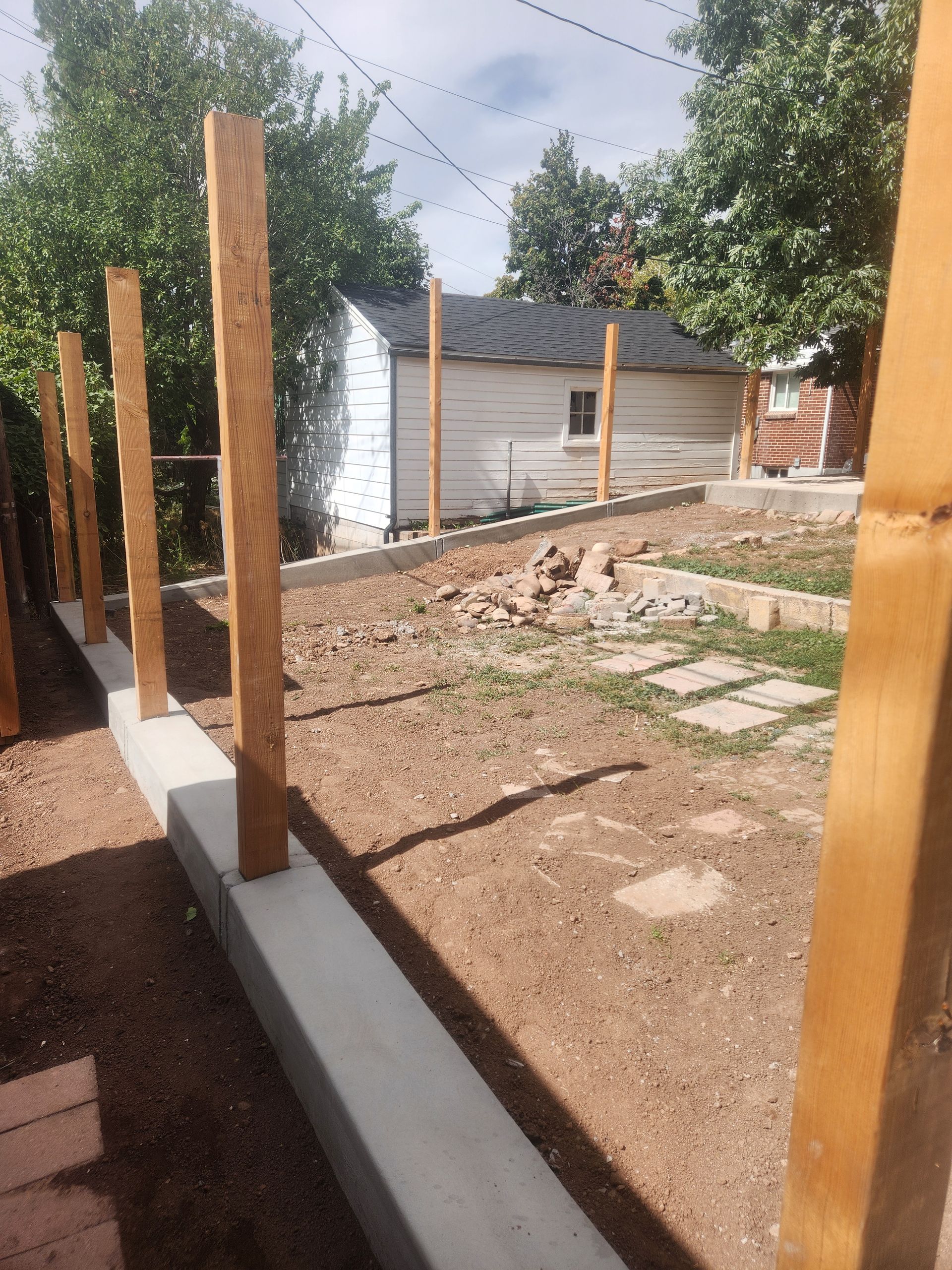 Wooden posts and a concrete foundation outline a yard under construction beside a small shed.