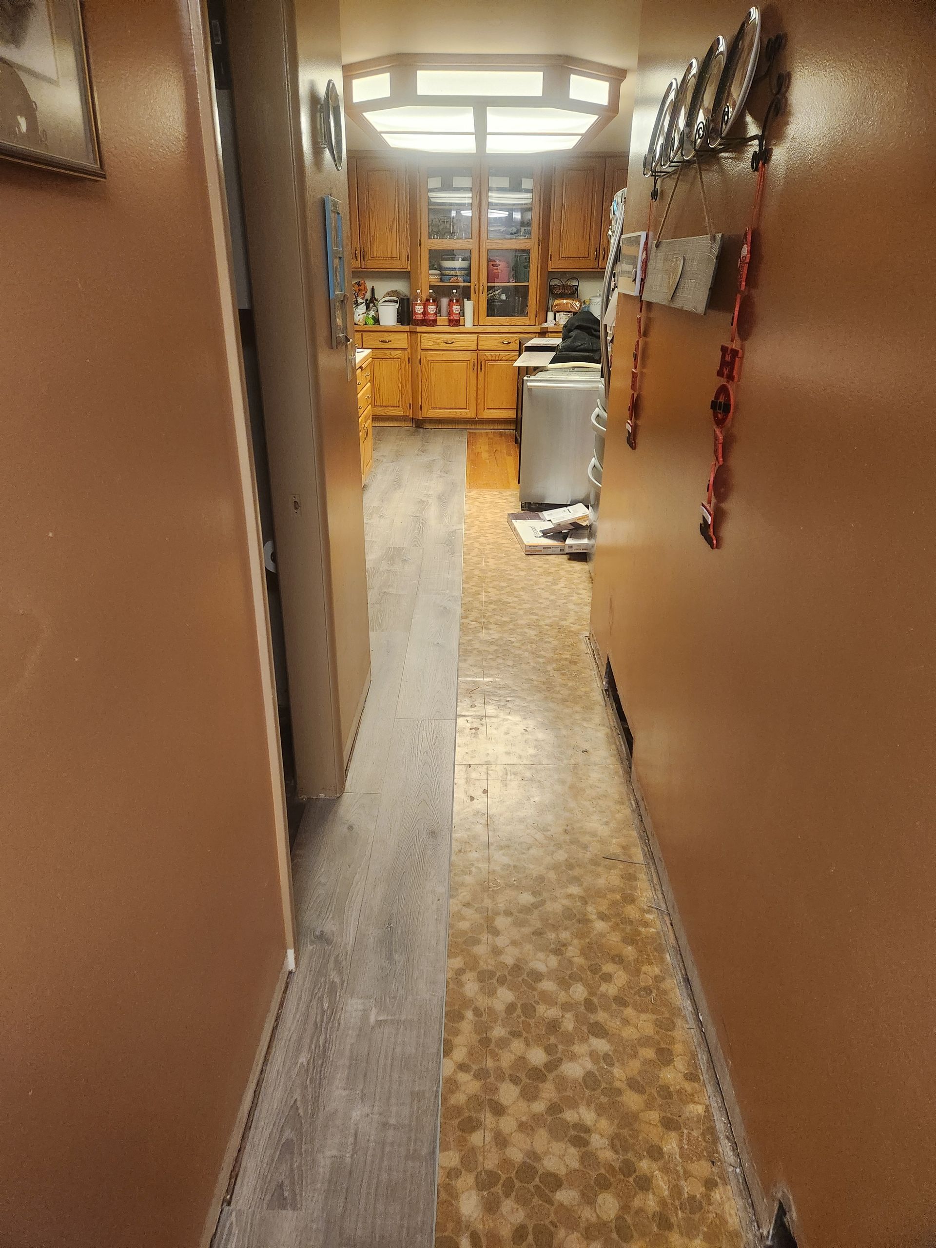 Narrow hallway with brown walls, leading to a kitchen or break room with wooden cabinets and a trash can.