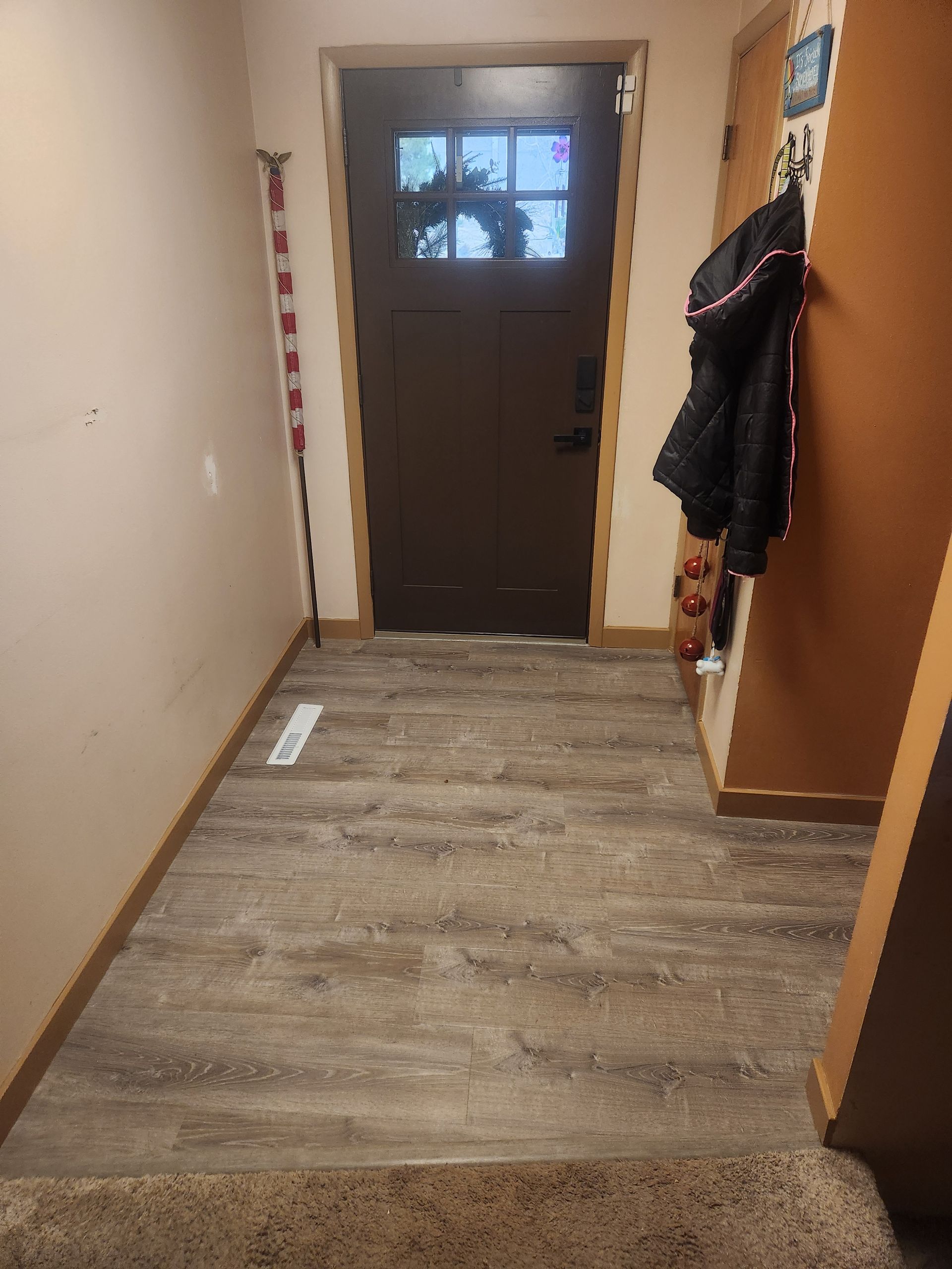 Narrow hallway with worn wood floor, brown front door, and coats hanging on the right.