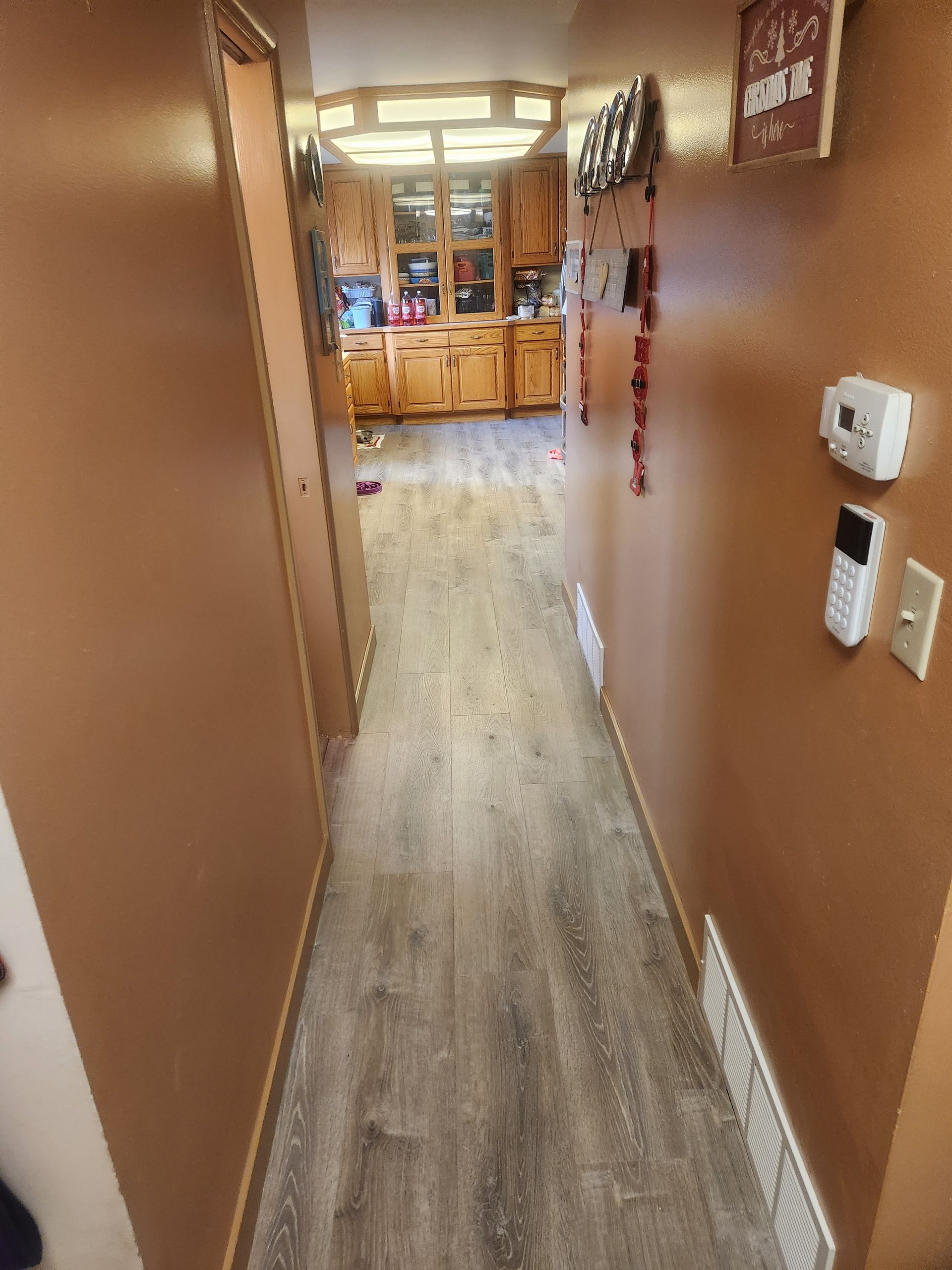 Narrow brown hallway with wood-look floor leading to a bright room with cabinets and a table.