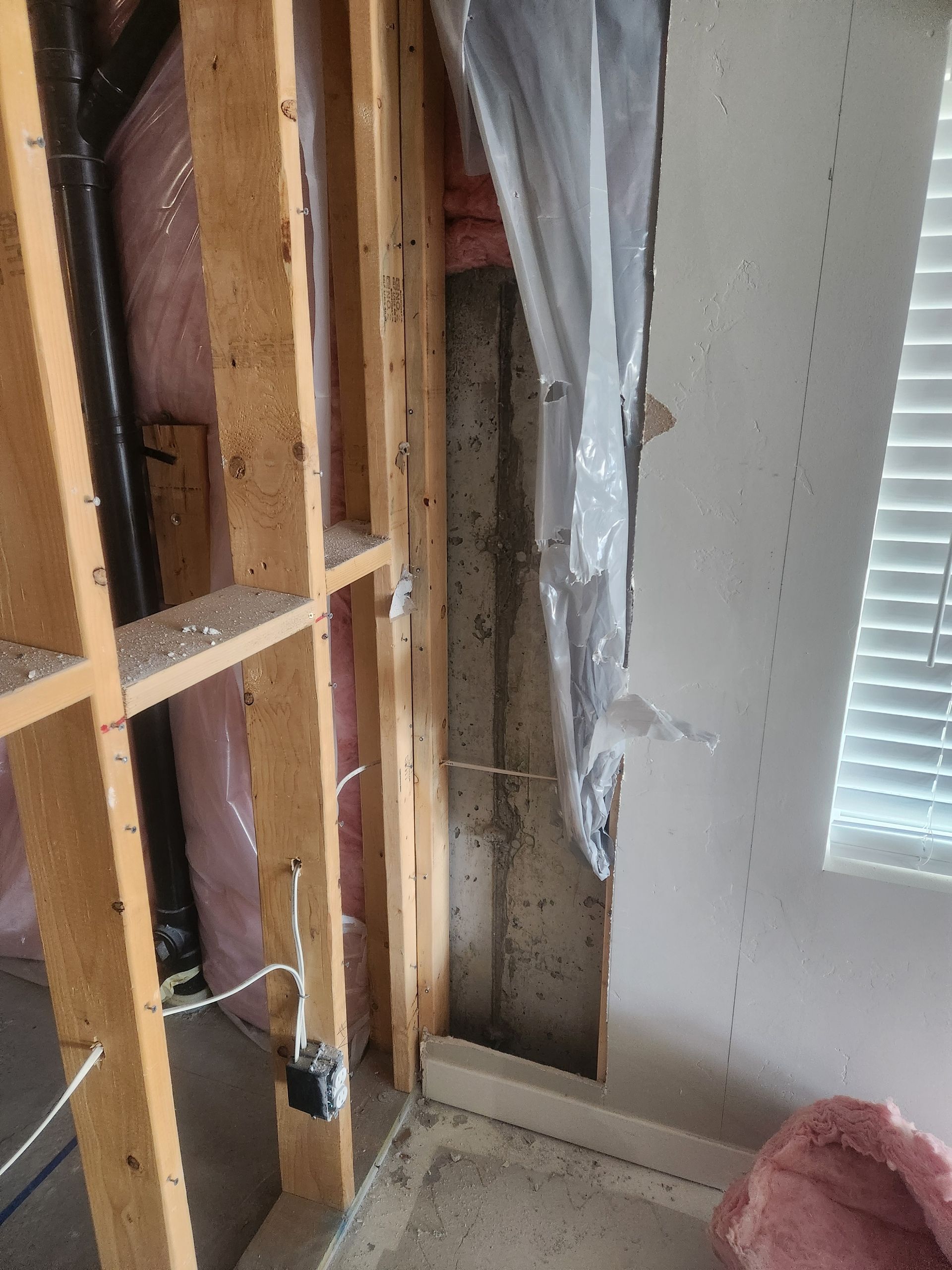 Exposed wall studs and wiring in an unfinished interior room beside a window with blinds