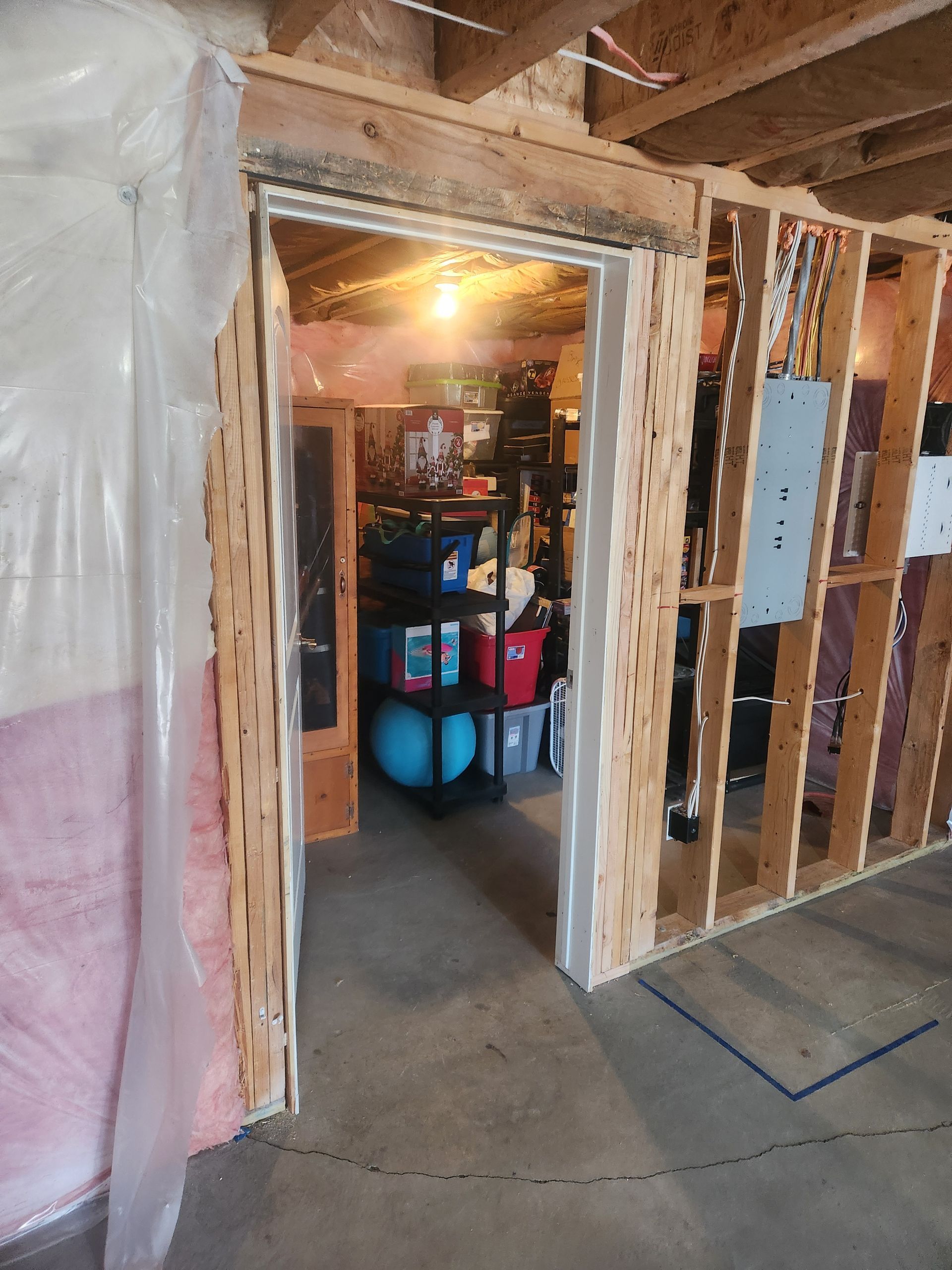 Unfinished basement doorway framed in studs and plastic sheeting, with storage bins visible inside.