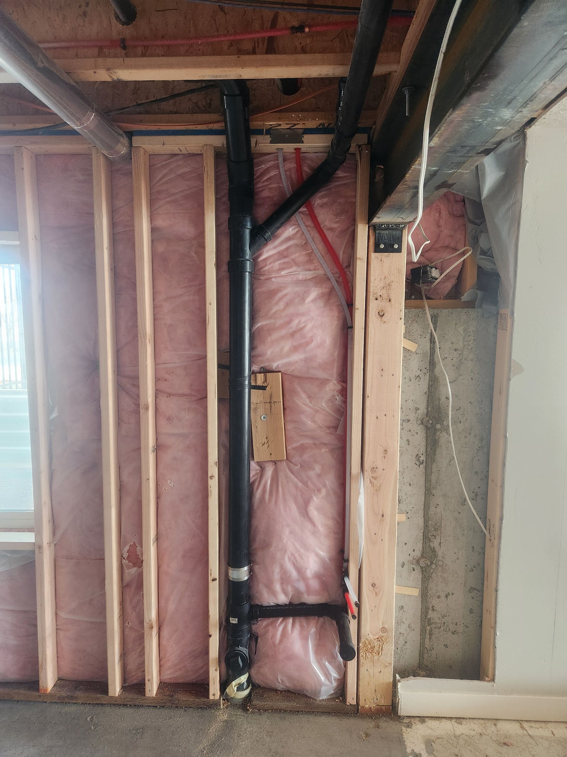 Exposed basement wall framing with pink insulation and black ducting beside a concrete wall.