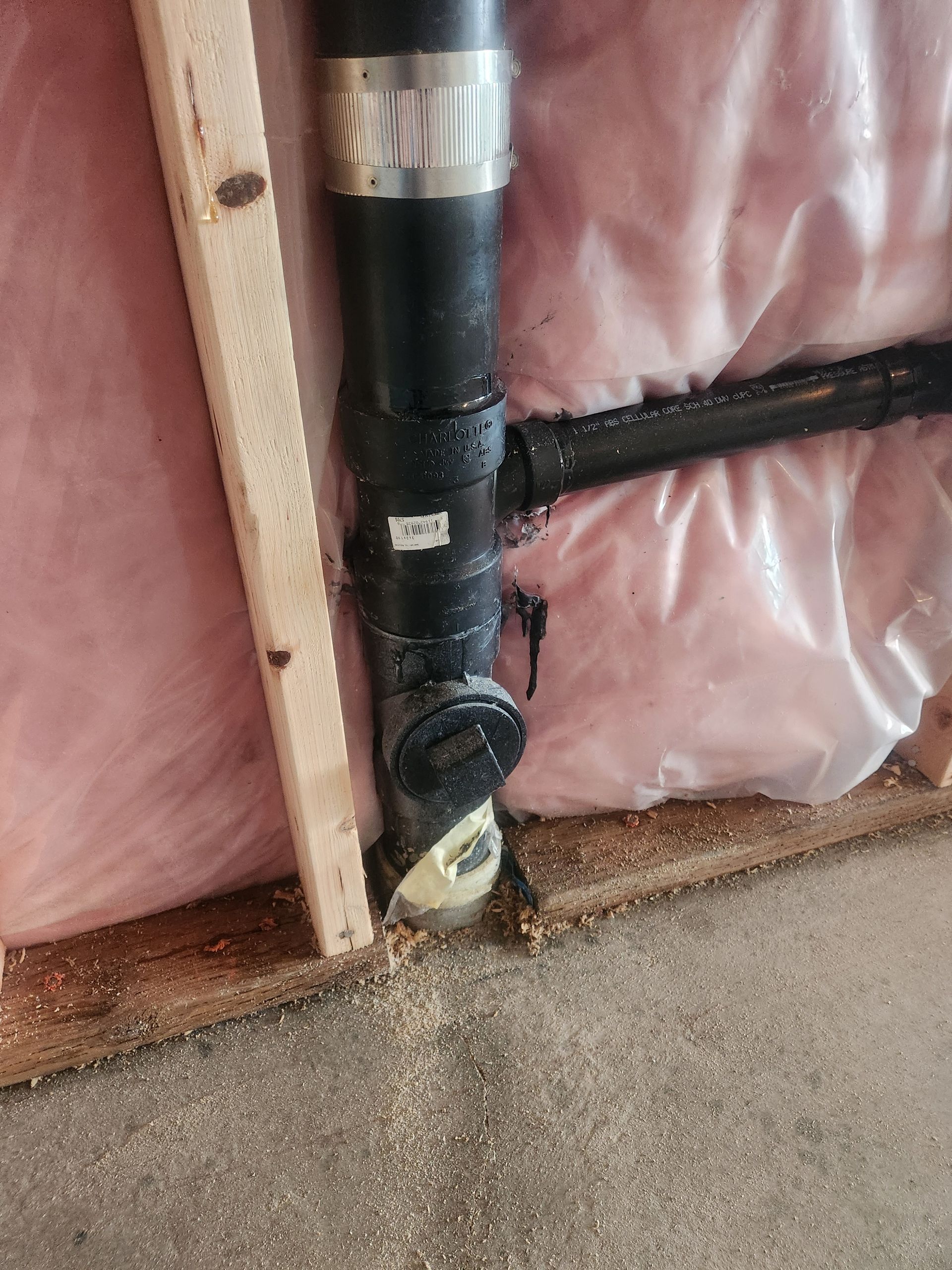 Black PVC pipe with white tape beside pink insulation and a wooden stud in a wall cavity