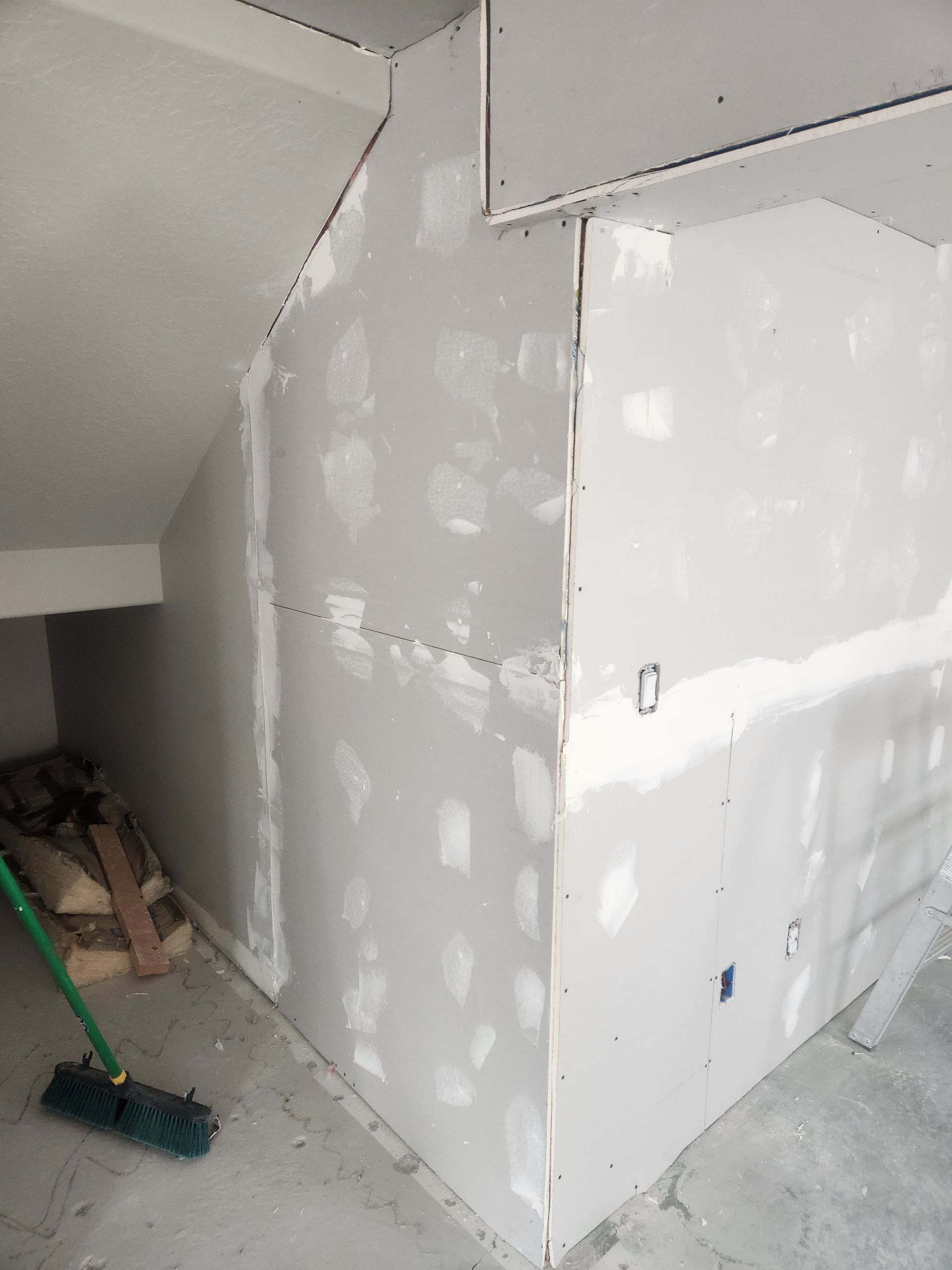 Partially finished drywall in an attic or storage space with patch spots and a broom on the floor