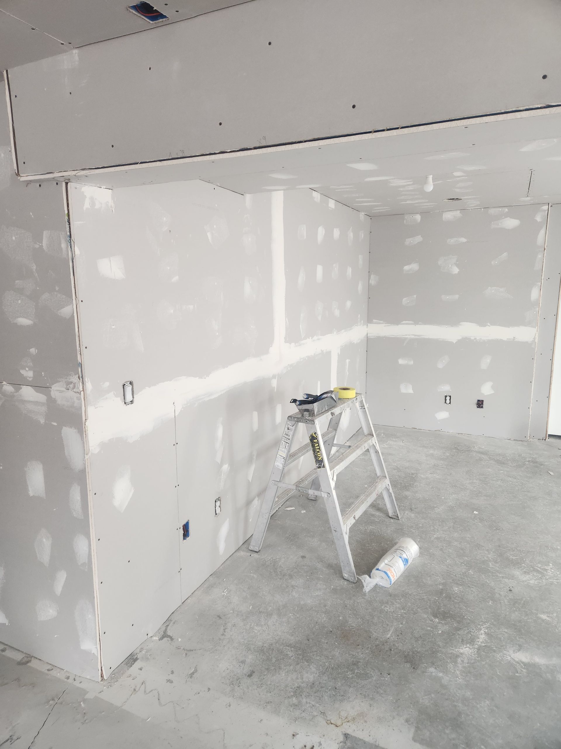 Drywall installation in an unfinished room, with a step ladder and tools on the floor.