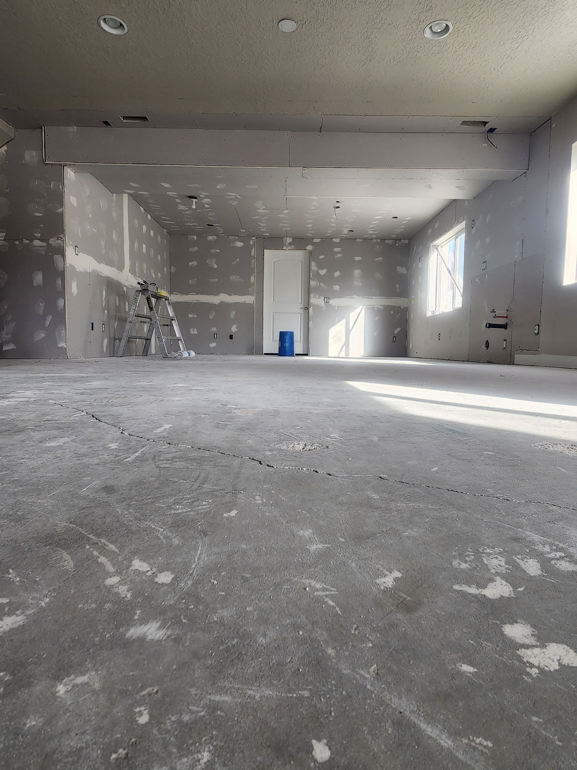 Empty unfinished room with gray drywall, concrete floor, and sunlight streaming through a window.