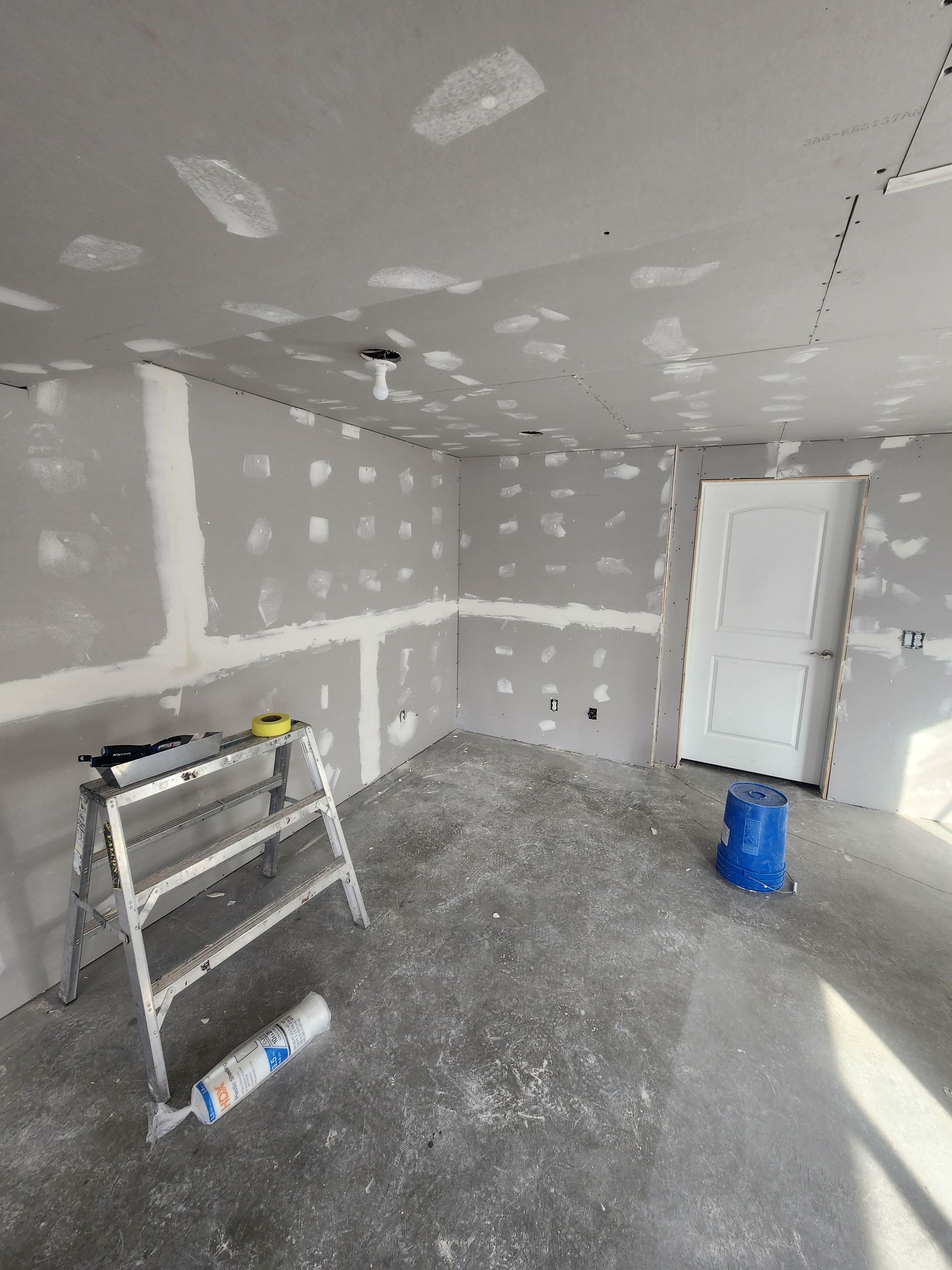 Unfinished drywall room with ladder, white door, and blue bucket on a concrete floor.