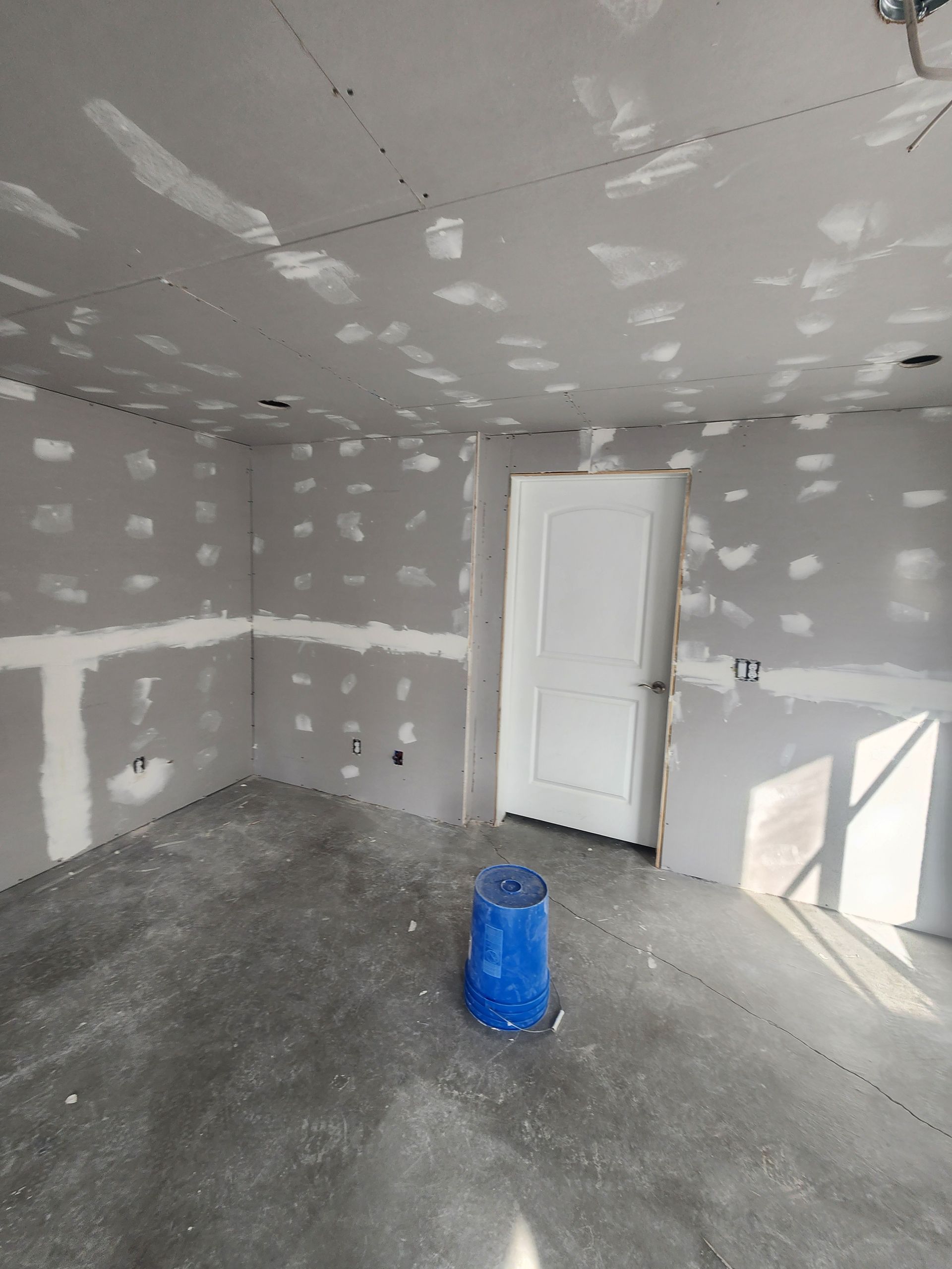 Empty unfinished room with gray drywall, a white door, and a blue bucket on the concrete floor.