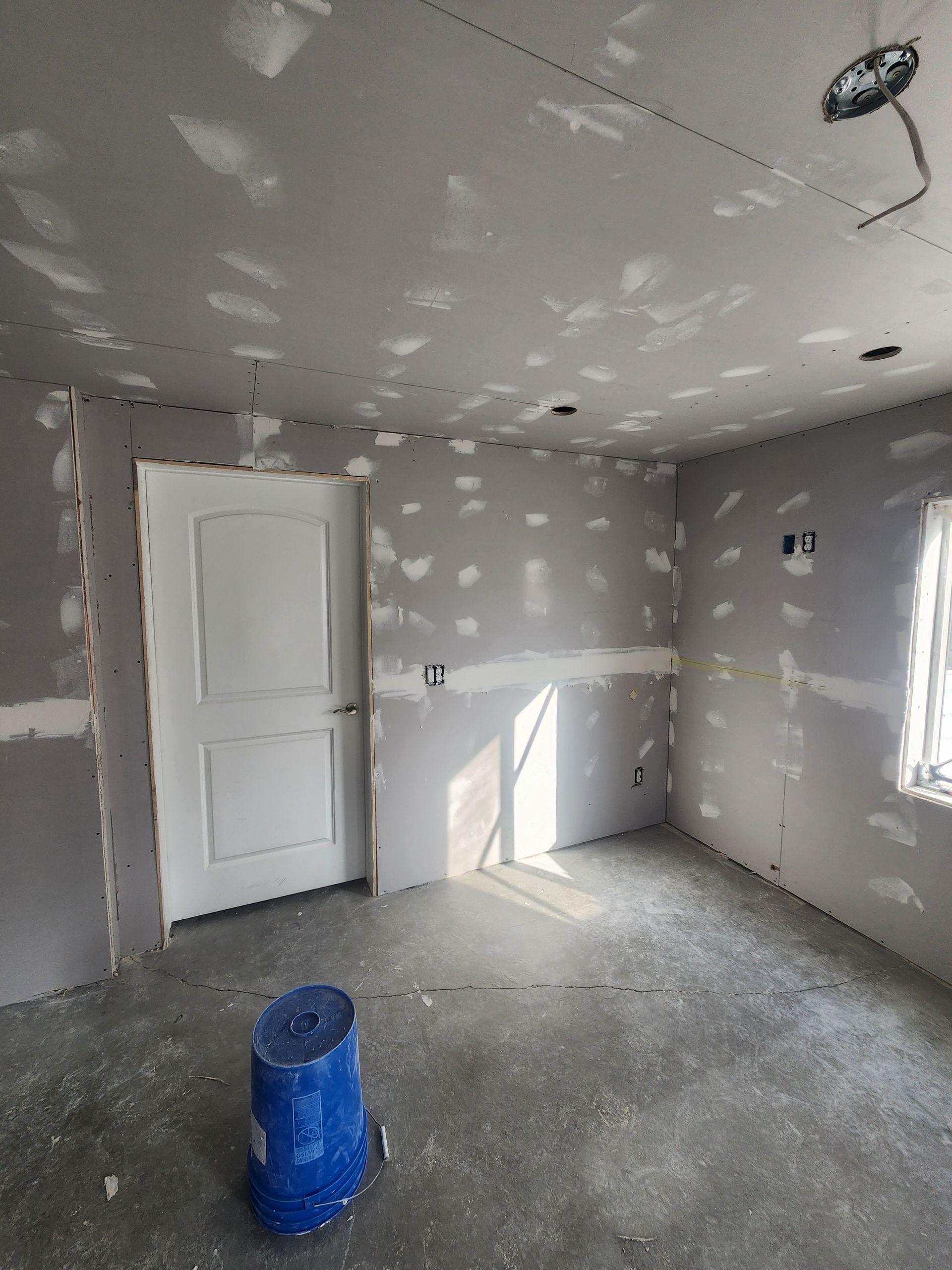 Unfinished room with gray drywall, white door, window light, and a blue paint bucket on the floor