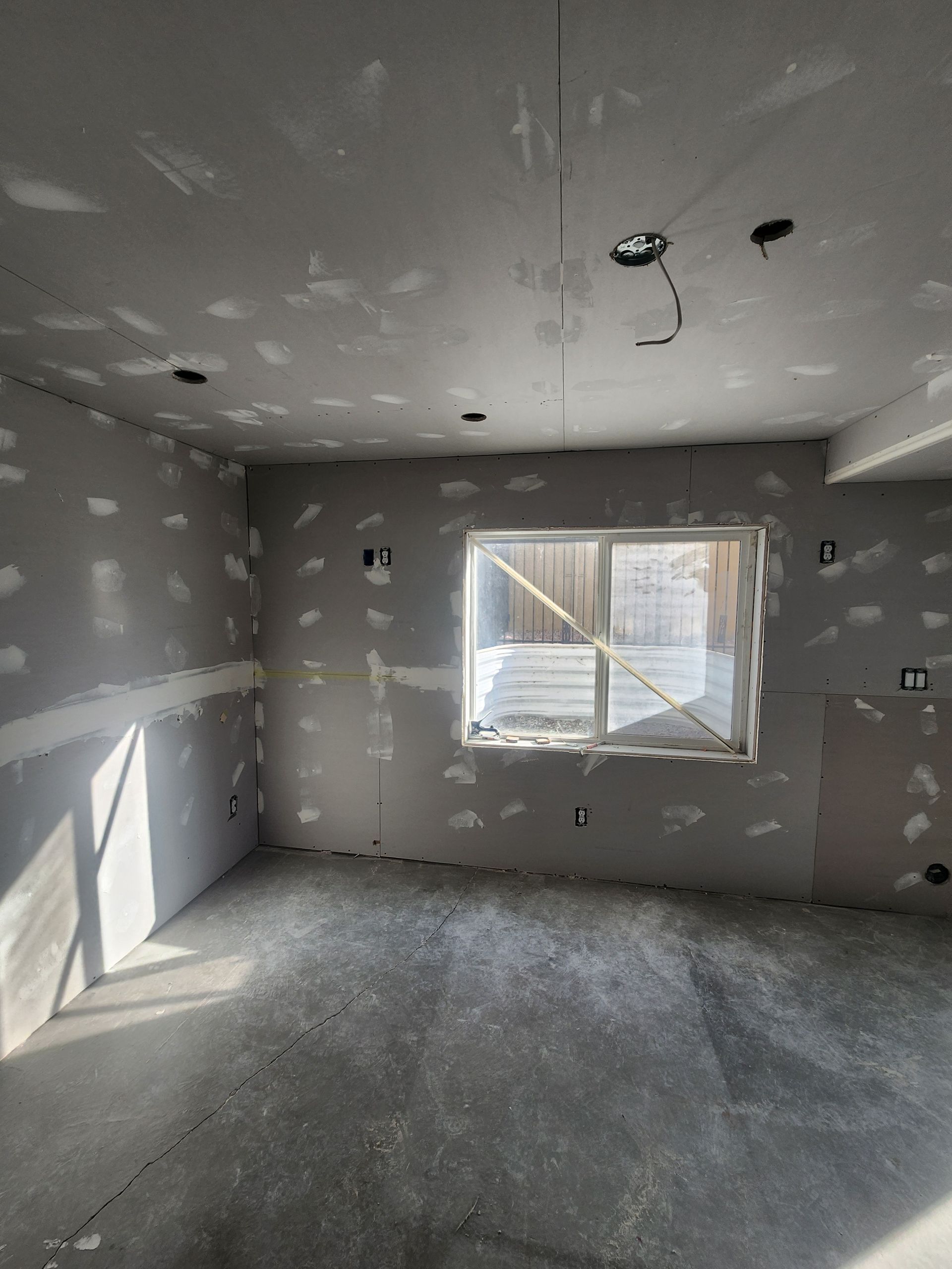Empty unfinished room with gray drywall, a window, ceiling light fixtures, and bare concrete floor.