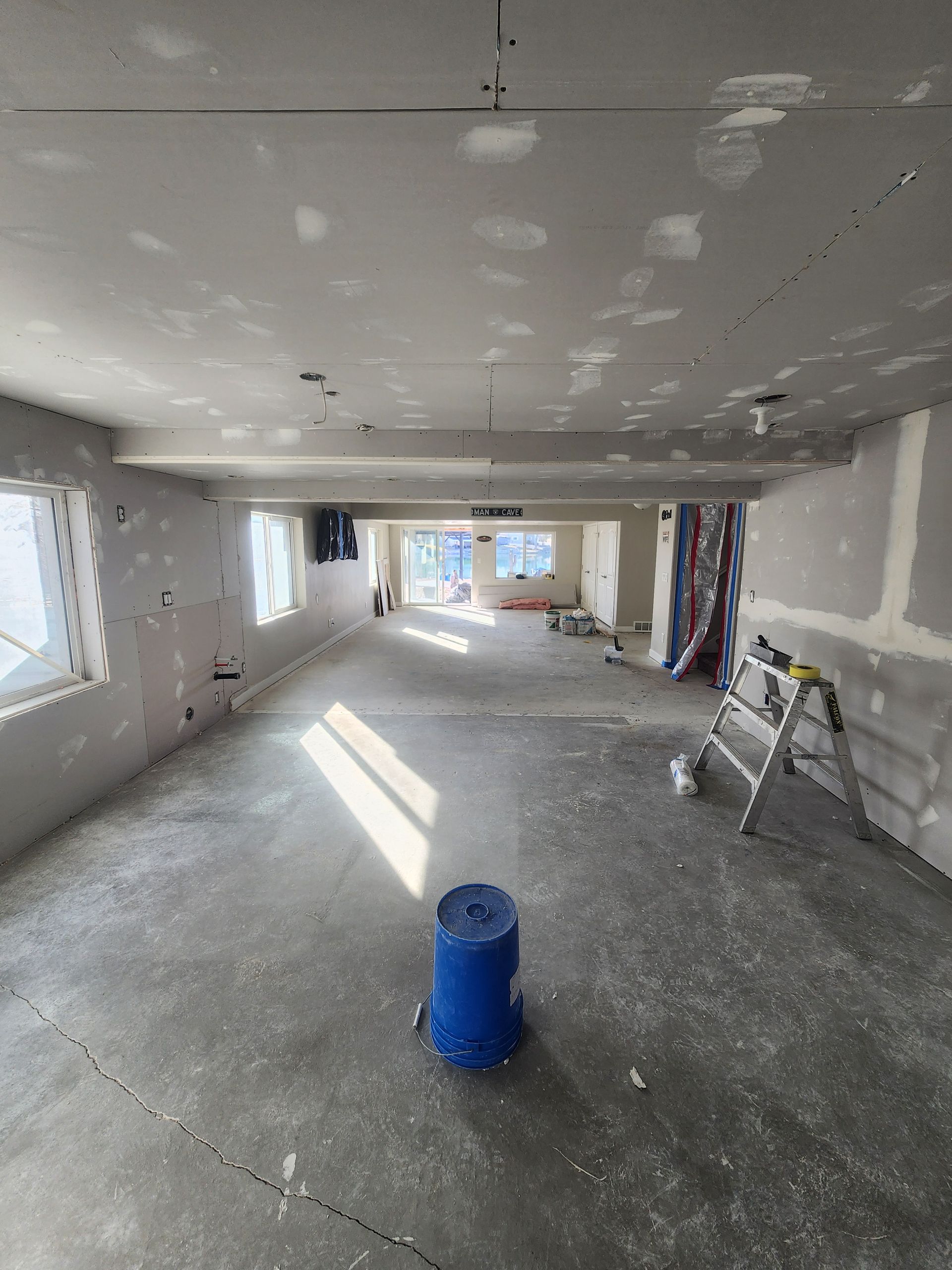 Unfinished interior room under renovation with drywall patches, windows, and a blue bucket on the floor