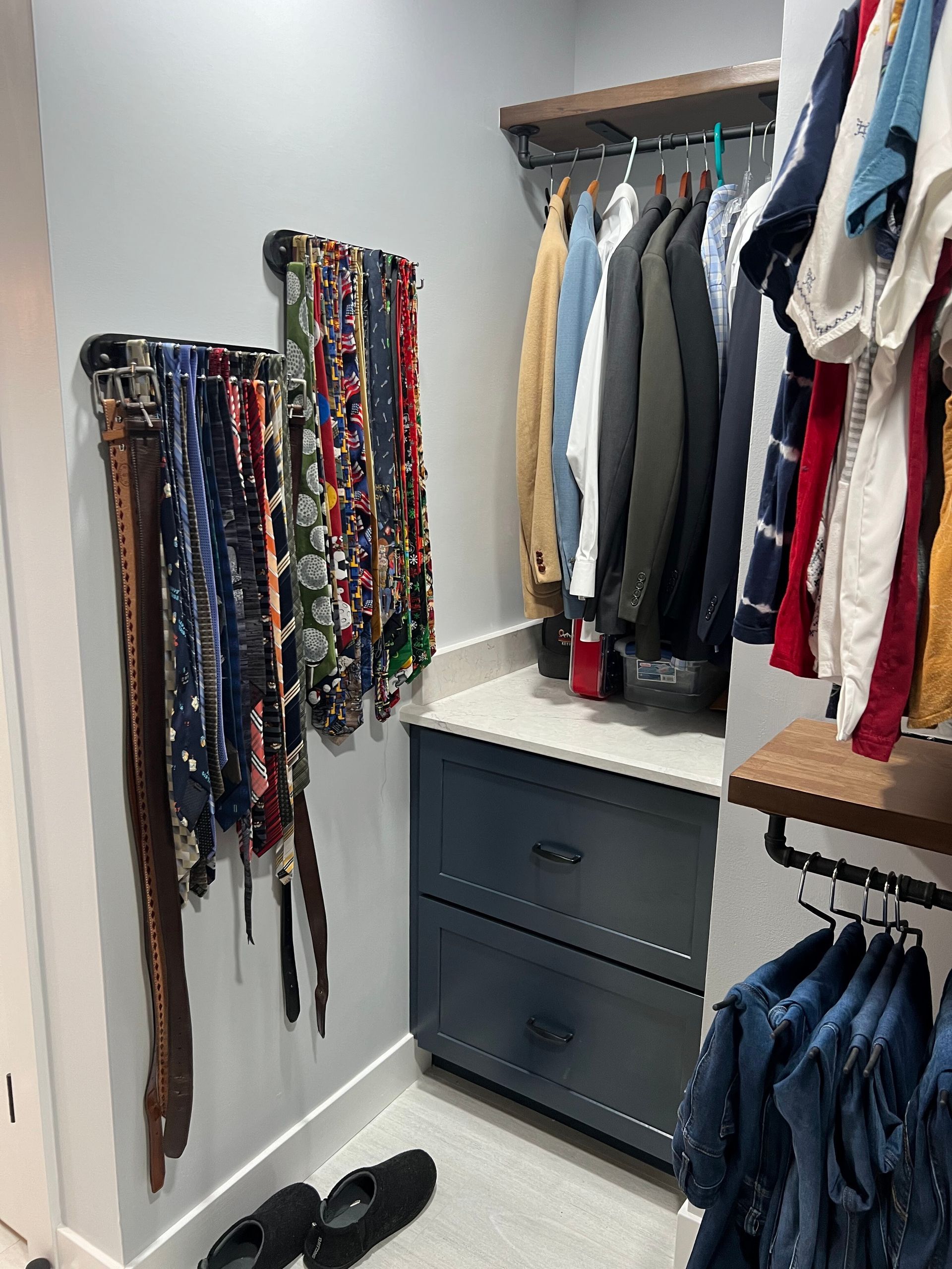 Walk-in closet with hanging clothes, belts on hooks, drawers, and shoes on the floor