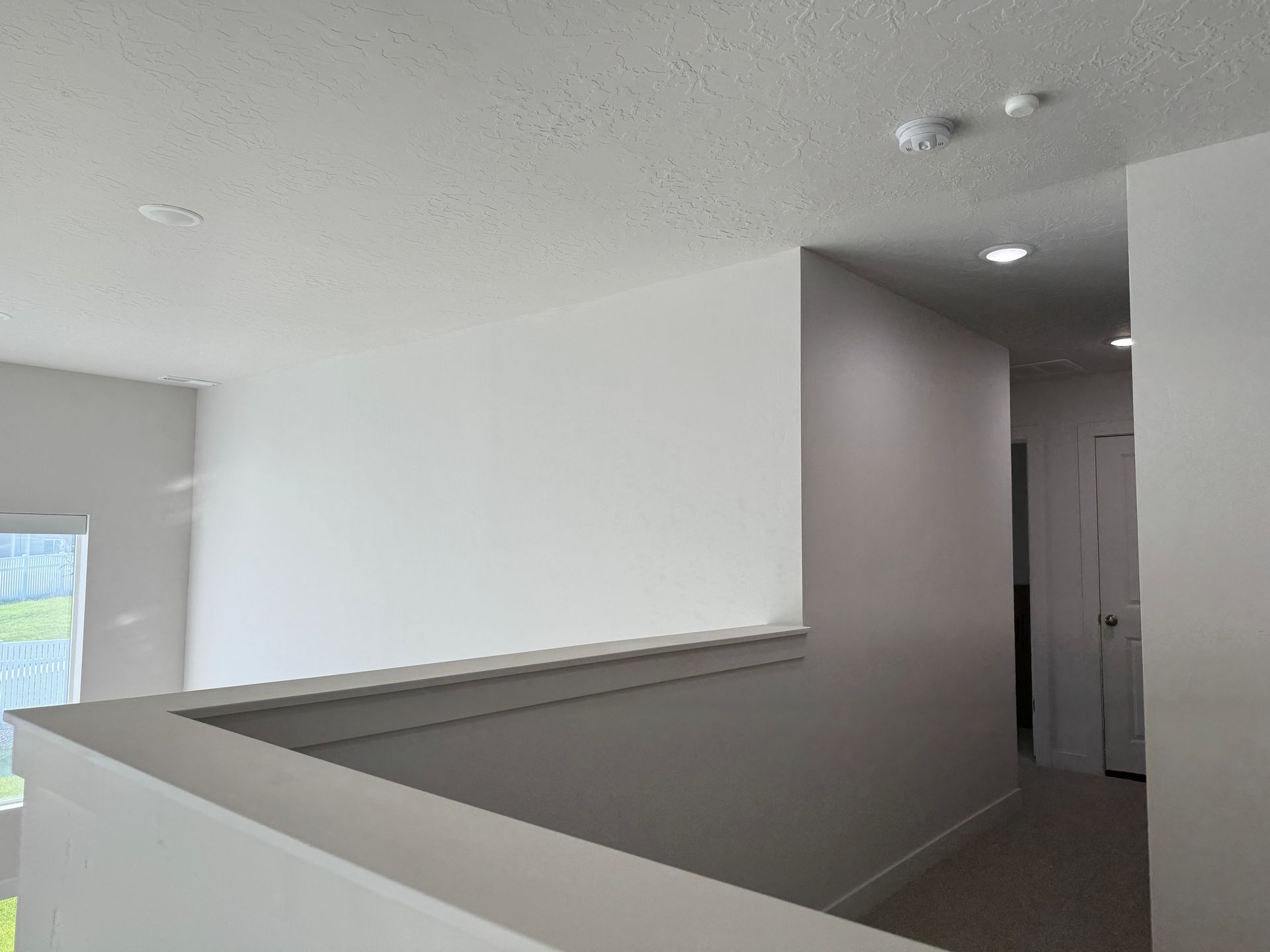 Interior hallway with white walls and ceiling, gray carpet, and a view into a small room.