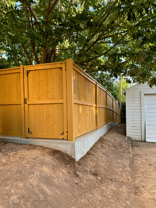 Yellow wooden fence beside a dirt path and white shed under leafy trees