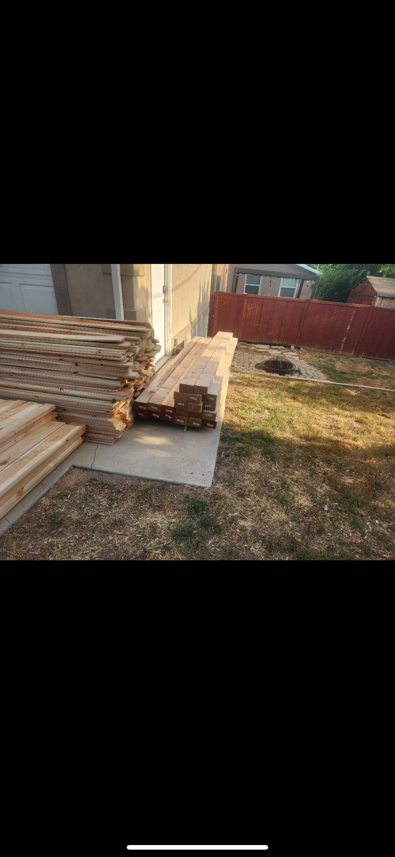 Stacked lumber on pallets beside a shed in a yard with dry grass and a red fence.
