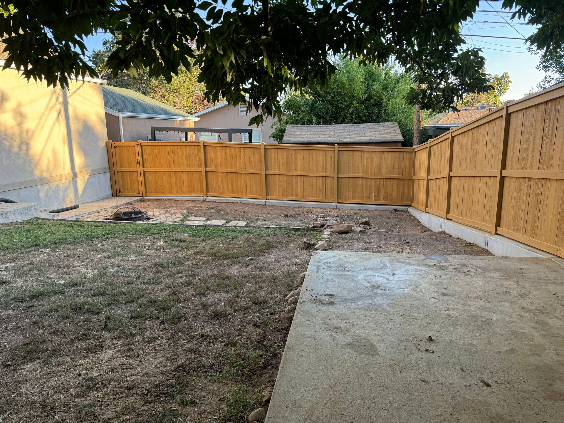 Backyard with a wooden fence, patchy grass, and a concrete patio under a tree canopy.