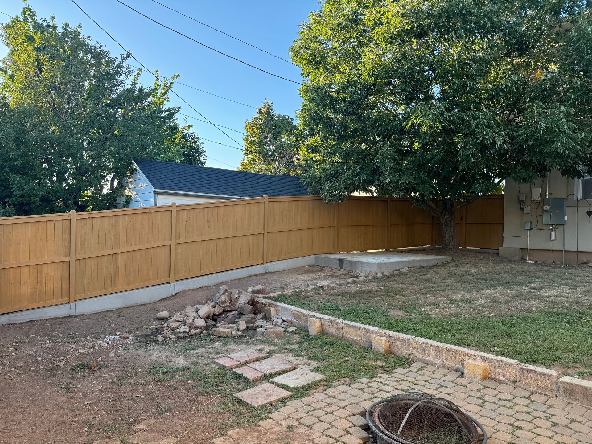 Backyard with a wooden fence, trees, patio pavers, and a small pile of rocks near a brick border