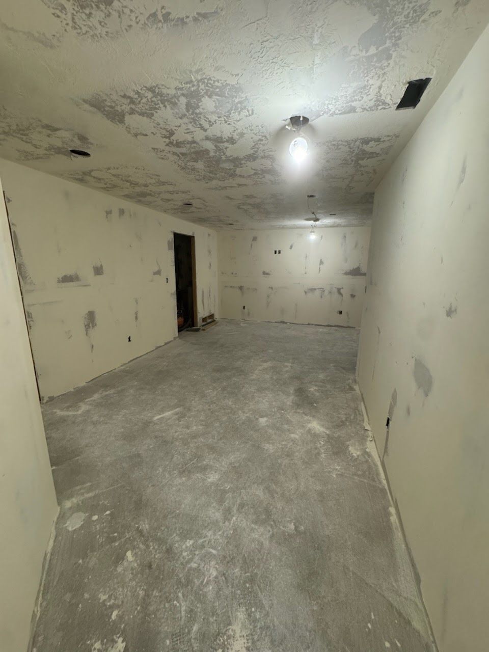 Empty unfinished room with bare concrete floor, stained walls, and a single exposed light bulb.