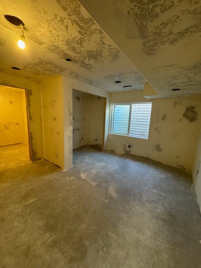 Empty, worn room with peeling ceiling paint, a window with blinds, and an open doorway lit by a yellow bulb