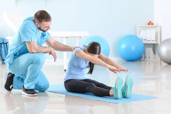 Fisioterapista assiste un paziente con esercizi di stretching da seduto in una luminosa sala di riabilitazione con palloni da ginnastica.