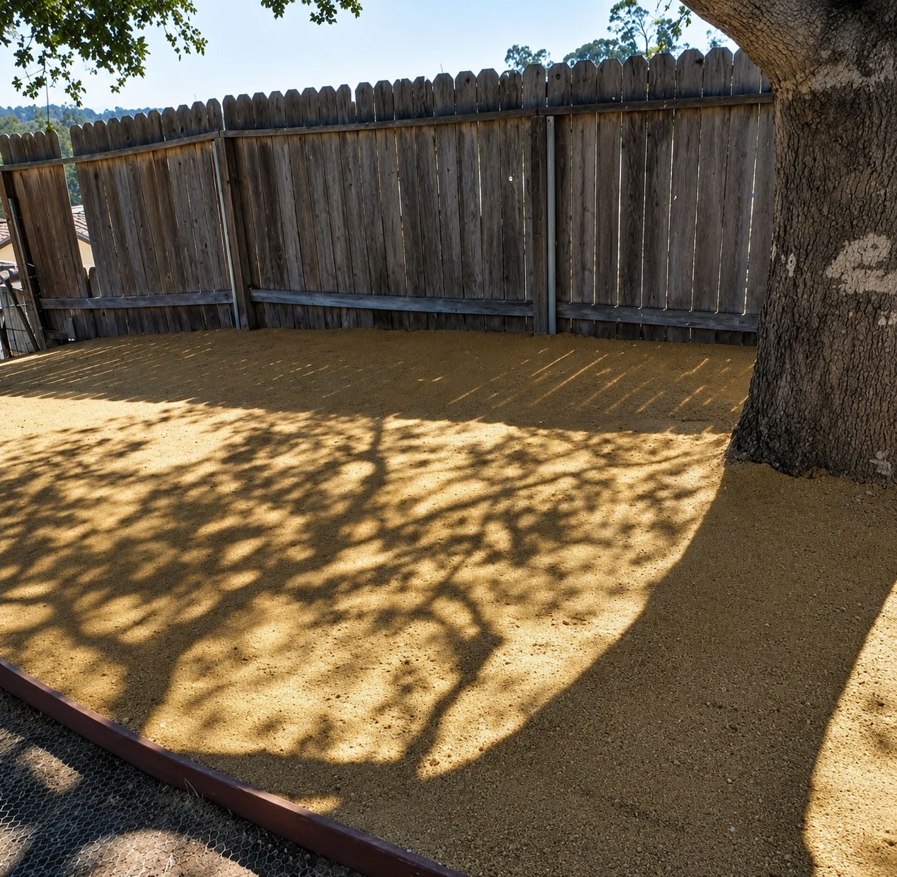 Backyard with a large tree casting shade on a gravel-covered area beside a wooden fence