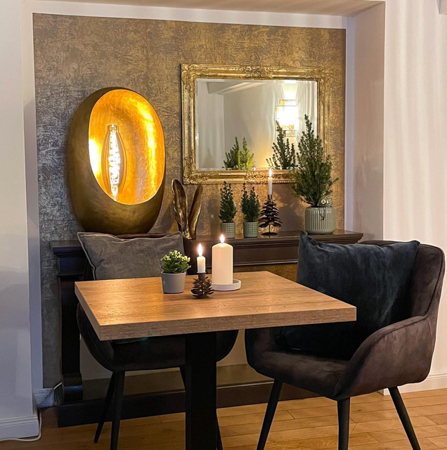 Vor einer goldbeleuchteten Lampe, einem Spiegel und zwei dunkelgrauen Stühlen steht ein kleiner Esstisch aus Holz mit Kerzen und einer Pflanze.