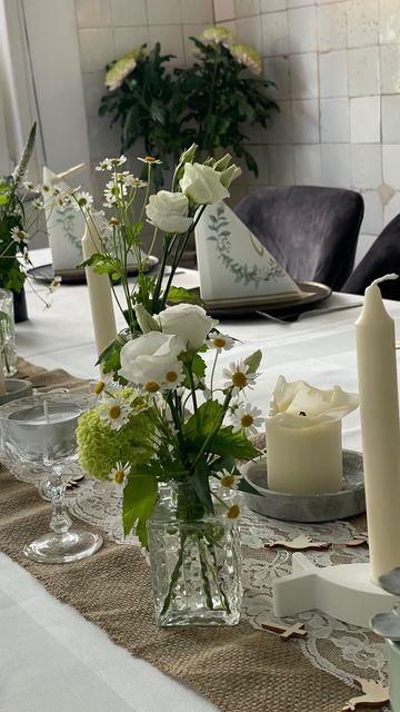 Ein für eine Feier gedeckter Esstisch mit weißen Blumen in Glasvasen, brennenden Kerzen und gefalteten Servietten auf Jute-Tischläufern.