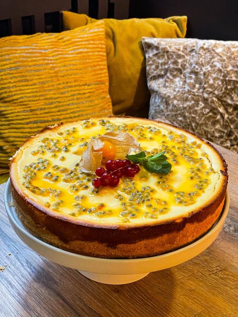 Ein mit Passionsfrucht, roten Johannisbeeren und einer Physalis garnierter Käsekuchen auf einem weißen Ständer mit verschwommenen Kissen im Hintergrund.
