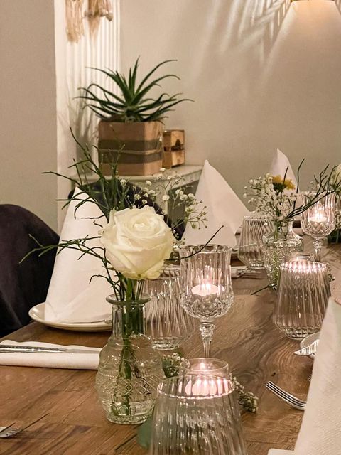 Ein rustikaler Holztisch, gedeckt zum Abendessen mit weißen Rosen in Glasvasen, brennenden Kerzen und gefalteten Servietten.