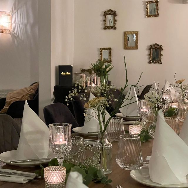 Ein formell gedeckter Esstisch mit gefalteten weißen Servietten, Kerzen und Blumen vor einer mit kleinen Spiegeln verzierten Wand.
