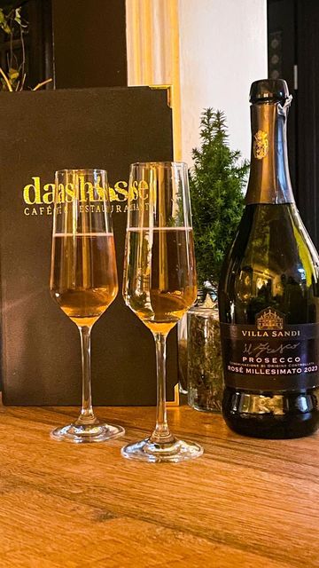 Auf einem Holztisch stehen zwei Gläser Sekt neben einer Flasche Champagner und einer schwarzen Speisekarte.