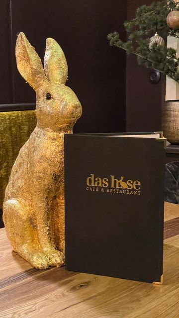 Auf einem Holztisch steht neben einer schwarzen Speisekarte mit goldenem Hasenlogo eine glitzernde goldene Hasenfigur.