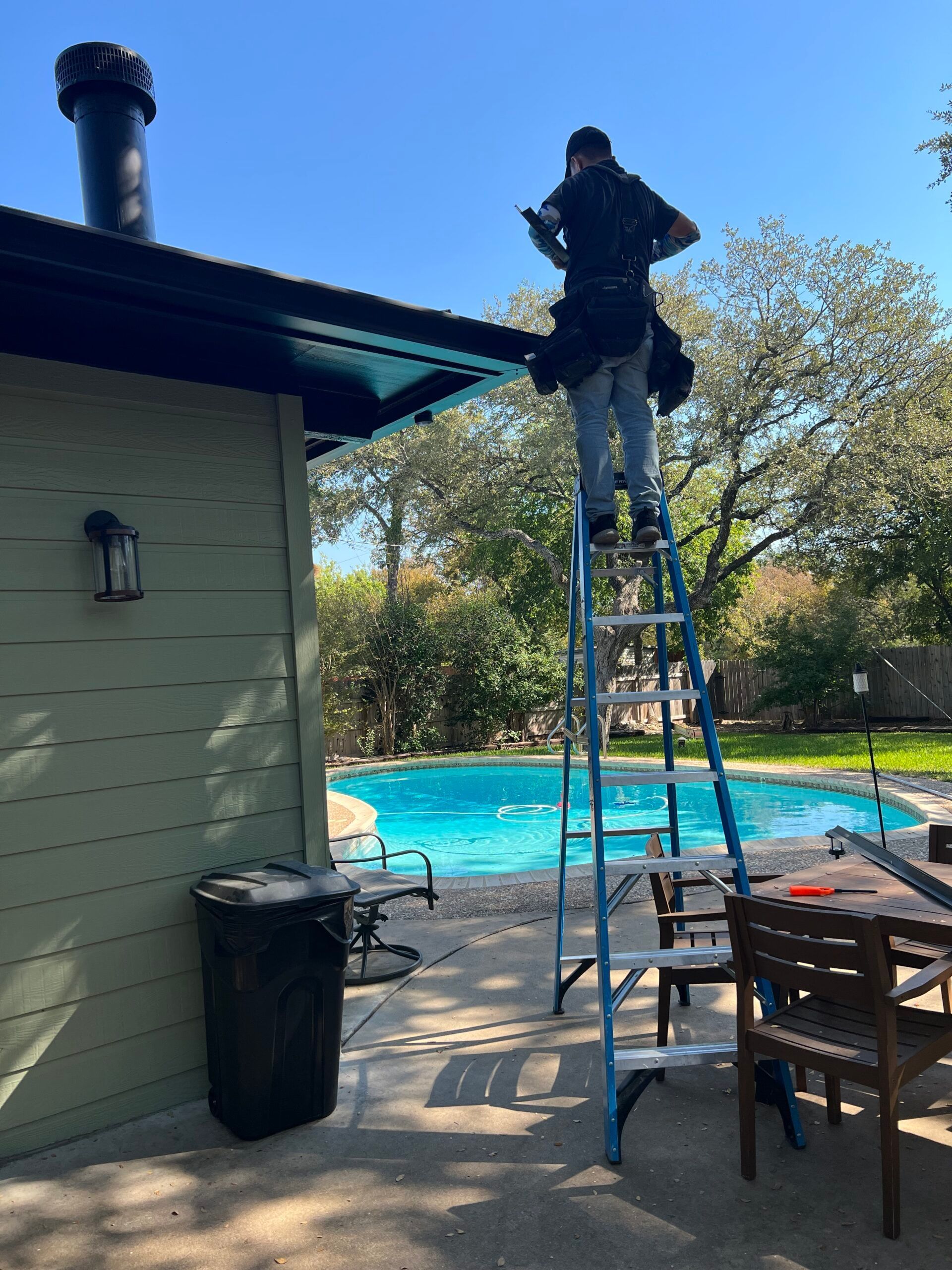 Person on a ladder cleaning gutters of a house with a pool in the background. Sunny day.