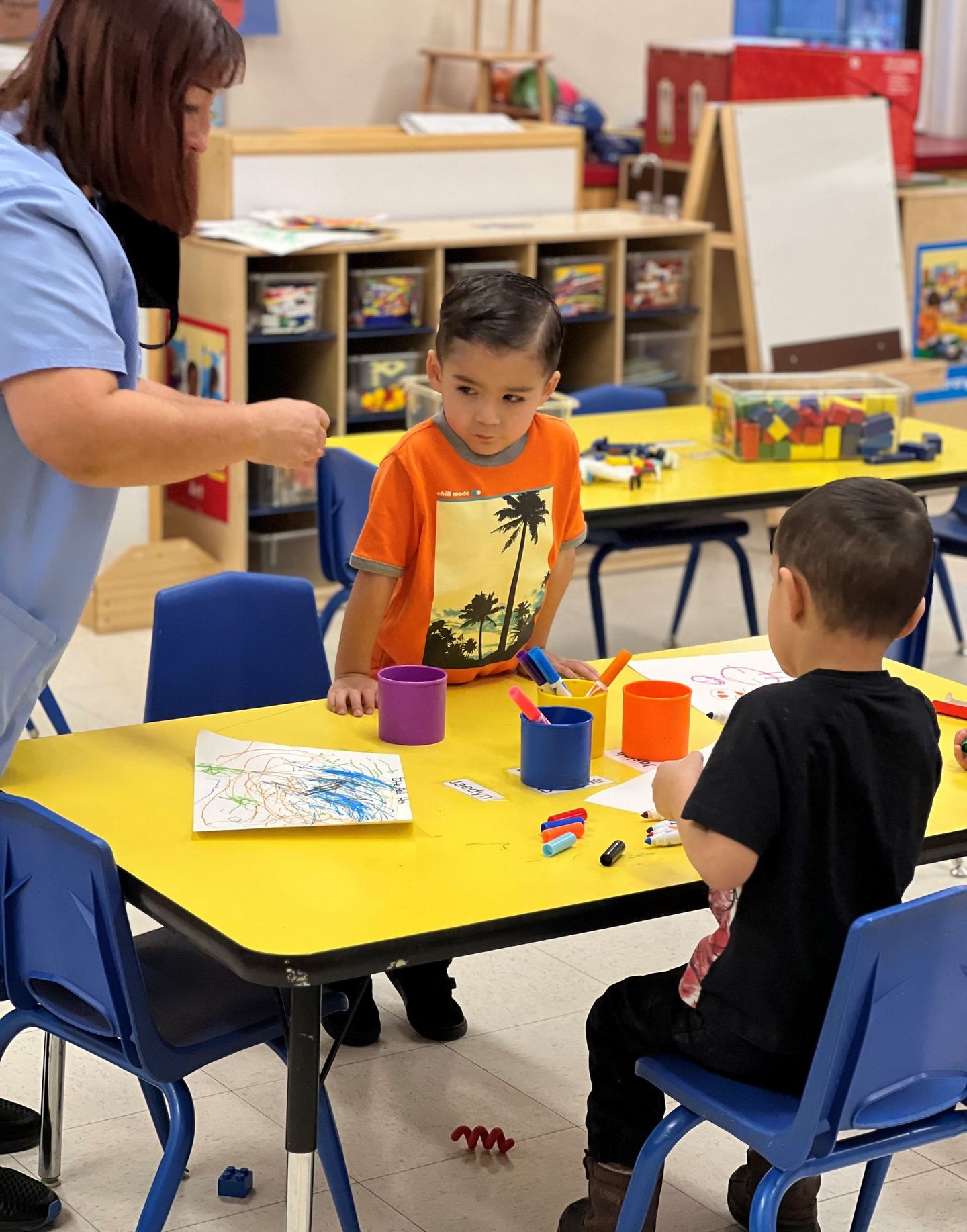 a woman is helping two young boys paint at a yellow table