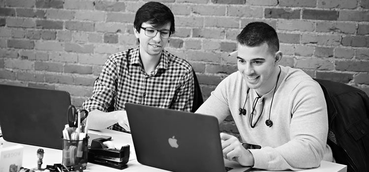 Two men are sitting at a table looking at a laptop computer.